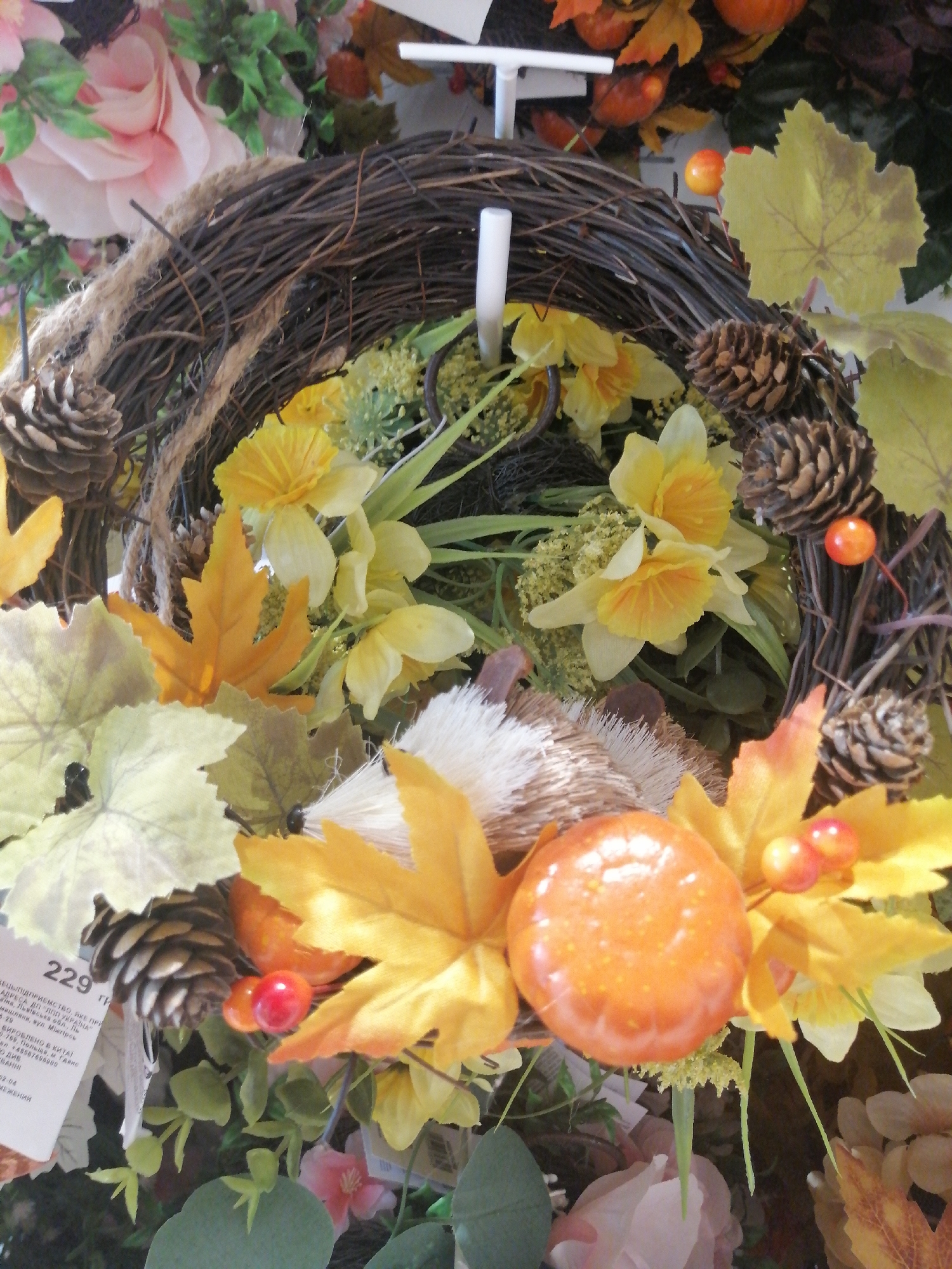The image shows a close-up view of a decorative fall-themed wreath. The wreath is constructed from twisted brown branches and is adorned with artificial yellow flowers, orange artificial berries, pinecones, and artificial autumn leaves in shades of yellow and orange. In the center of the wreath, there is an artificial orange mushroom or pumpkin-shaped ornament. A white price tag with Cyrillic text, indicating it is likely in Kremenchuk, Ukraine, is attached to the wreath. The background is blurred but suggests a shop or market setting with more floral decorations. There are no people visible in the image.