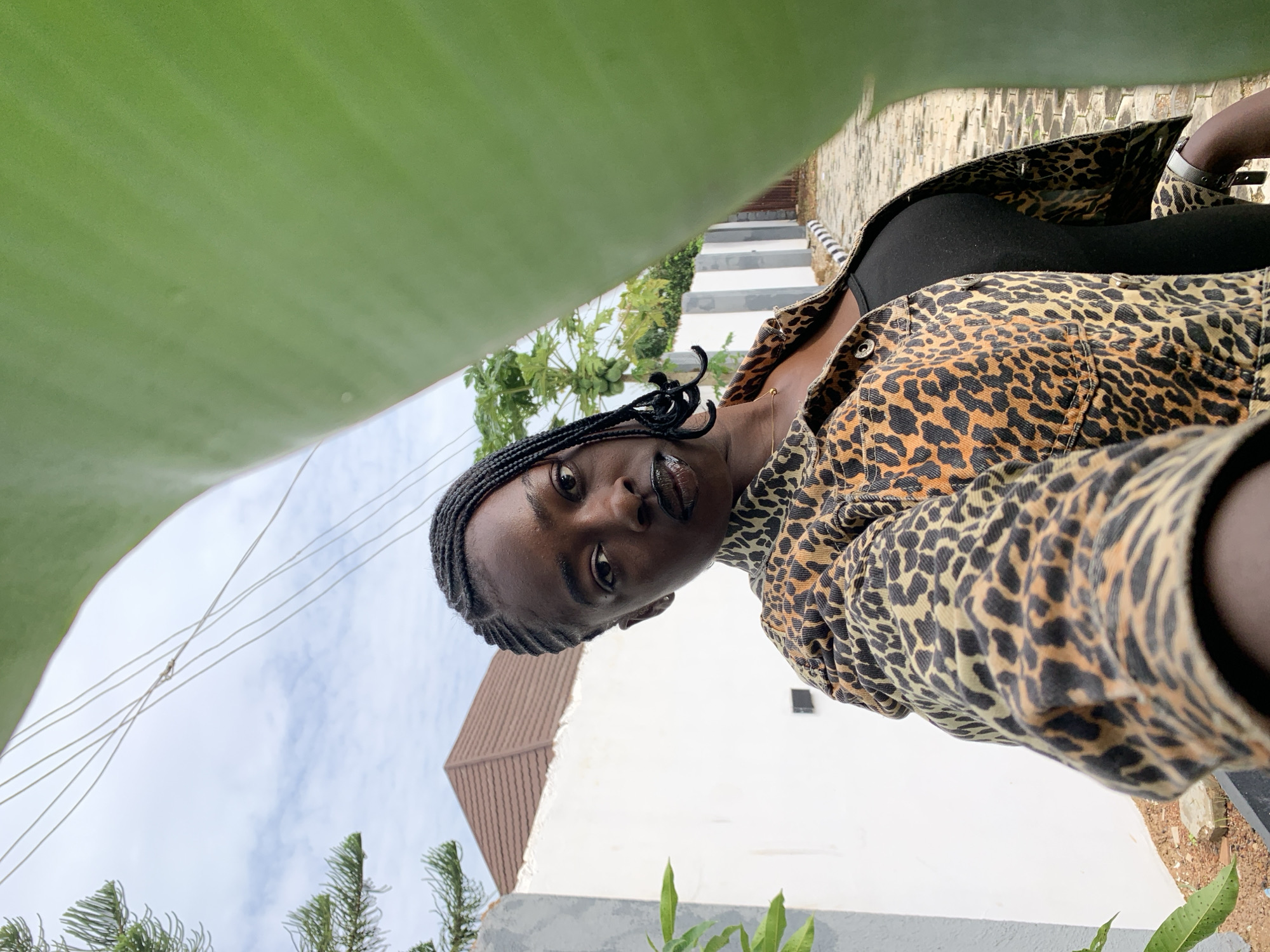 A young Black woman with dark skin and braided hair is taking a selfie outdoors, framed by a large green leaf. She is wearing a leopard print denim jacket over a black top, with dark lipstick. In the background, there is a white wall, a brown roof, a tree with green fruit, and a paved walkway with black and white striped curbs. The sky is cloudy. The location is the city of Gura Top, Nigeria.