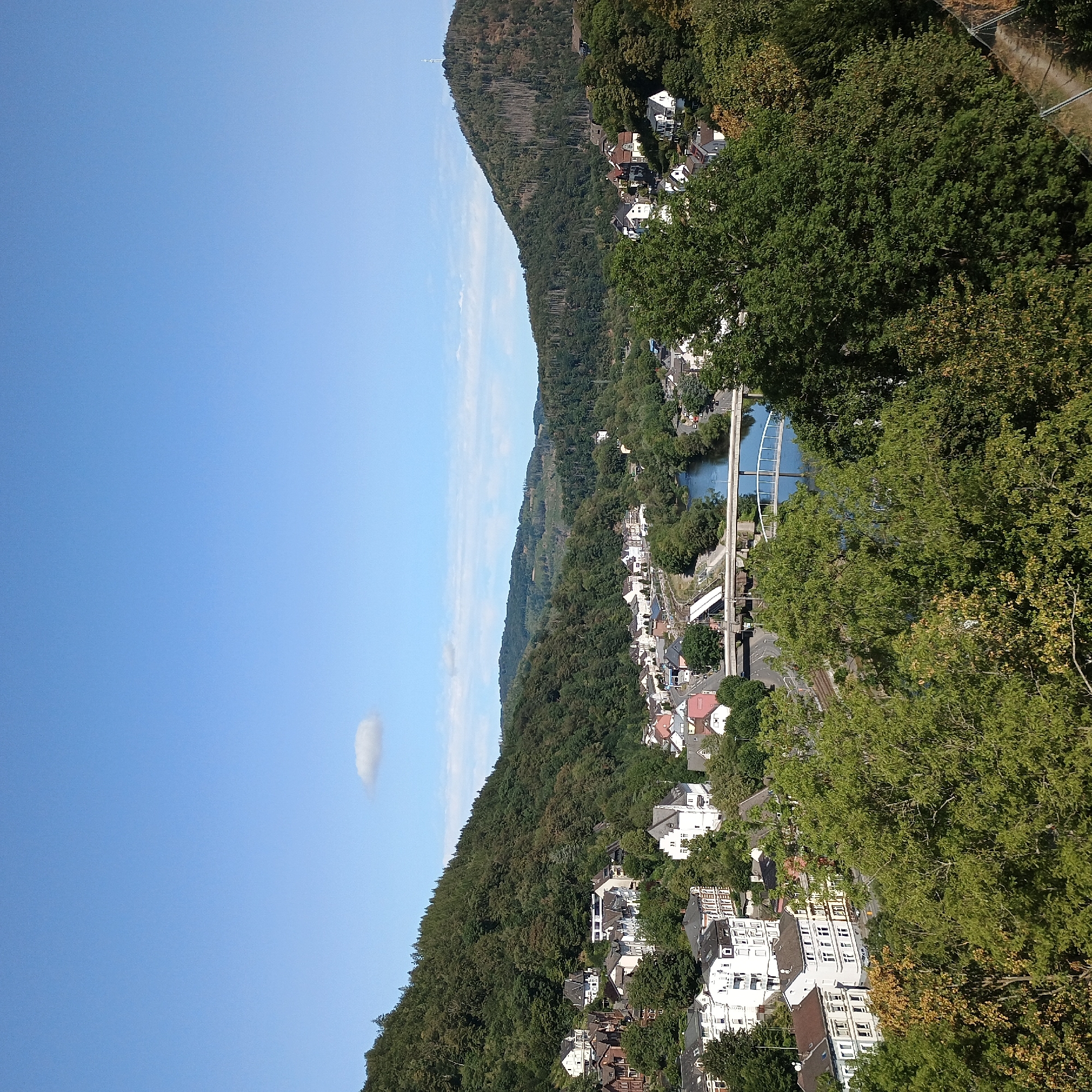 This is a high-angle, wide shot of a town nestled in a valley, with steep, tree-covered hills on either side. The town features a cluster of white and brown buildings with red roofs. A river runs through the town, crossed by at least two bridges. The sky is clear blue with a single, small white cloud visible. The foreground is dominated by dense green trees. The location is identified as Altena, Germany.