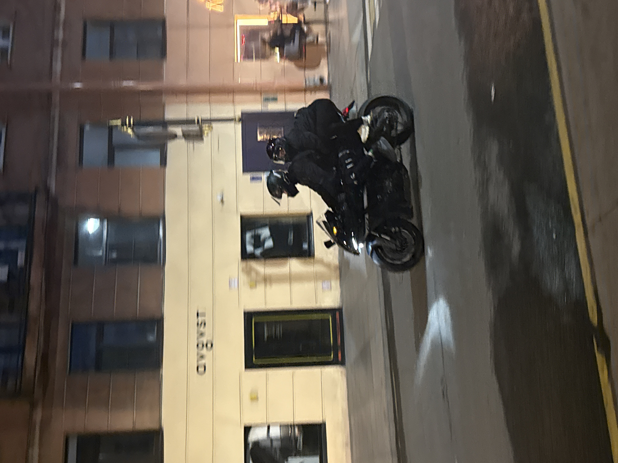 The image captures a nighttime street scene in Saint Petersburg, Russia, with a focus on two individuals riding a dark-colored motorcycle. The motorcycle is in motion, and the riders are wearing helmets and dark clothing, obscuring their identities. The background features a building with multiple windows and a sign that appears to read "OWOST" in stylized lettering. The street surface is wet, indicating recent rain or damp conditions, and there are reflections of lights on the ground. The overall lighting suggests it is evening or night.