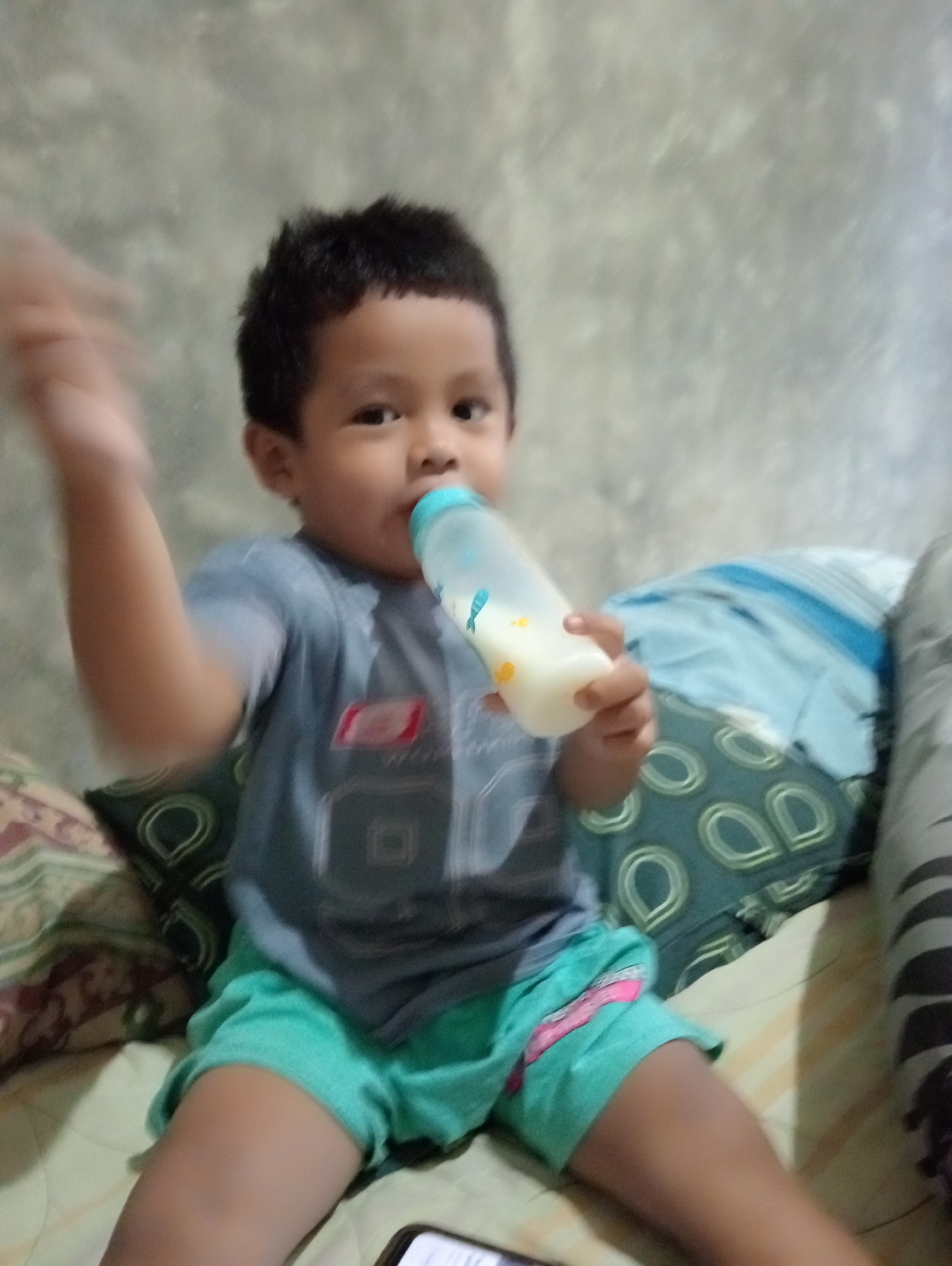 A young boy, appearing to be of Indonesian descent, is sitting on a bed while holding a baby bottle and drinking from it. He is wearing a gray t-shirt with a graphic design and teal shorts. The bed is covered with a patterned blanket. The background shows a textured wall. The image was taken in Muara, Indonesia.
