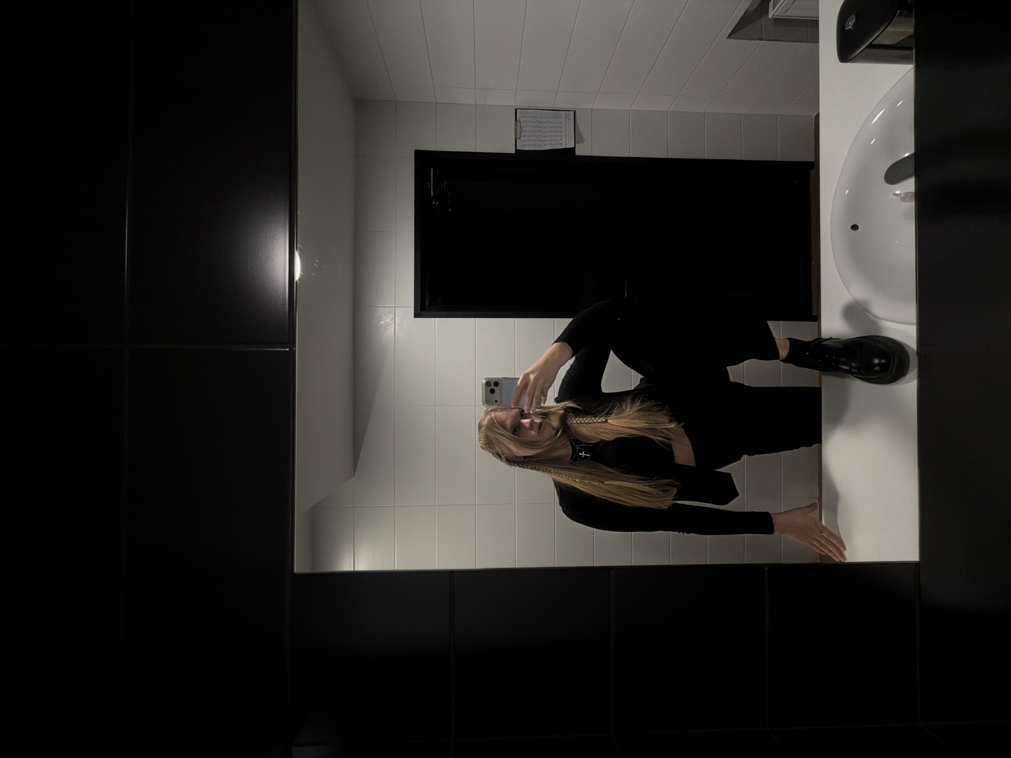 A young woman with long, straight blonde hair, styled with two prominent braids, takes a mirror selfie in what appears to be a modern restroom or changing room in Izhevsk, Russia. She is holding a light-colored smartphone, visible in the reflection, to capture the image. Her attire consists of a long-sleeved black top and black trousers, complemented by dark, possibly platform, shoes. A necklace with a black cross pendant is visible around her neck.
The setting is characterized by a high contrast monochromatic aesthetic. The walls are primarily white tiled, with a lower section of black tiling. The ceiling is also tiled in white. On the right side of the frame, a white sink with a dark faucet and a dark object, possibly a paper towel dispenser or hand dryer, are visible. In the background, above a dark door or window frame, a piece of paper, possibly sheet music or notes, is clipped to a board on the white tiled wall. The lighting appears to be artificial, typical of an indoor public space. The woman strikes a casual pose, with one arm extended towards the sink counter. The image is oriented with the woman's head towards the top left corner, indicating it has been rotated 90 degrees counter-clockwise from its original capture.