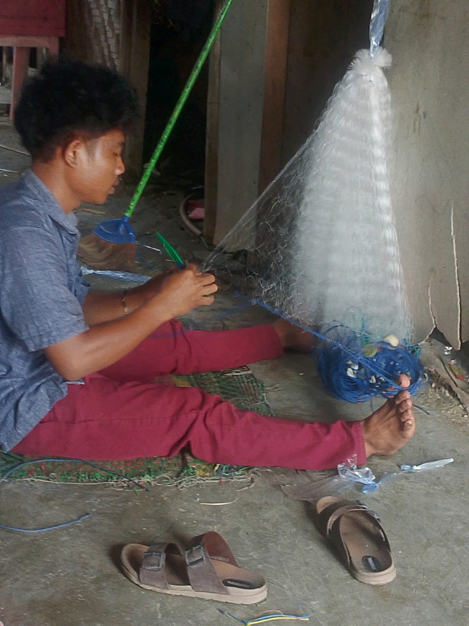 An Indonesian man, seated on the ground in what appears to be a rural or semi-rural environment in Labuhan Maringgai, Indonesia, is actively engaged in repairing or constructing a fishing net. He is wearing a short-sleeved grey collared shirt and maroon pants. His legs are extended in front of him, and his feet are bare. The fishing net, primarily white and with a wide opening, is suspended vertically and appears to be attached to a pole held by the man. A coil of blue rope, likely used for net construction or repair, rests beside the net, with several small floats visible within it. A broom with a blue dustpan attachment lies on the ground near the man's left side. The ground surface is concrete or packed earth, with some debris and loose strands of material scattered around. Two pairs of sandals are also present on the ground: one pair of brown, buckled sandals to the right of the man, and another lighter-colored pair of sandals further to the left. The background suggests an outdoor or semi-enclosed structure, possibly a workshop or storage area, with a wall visible behind the net. The overall scene depicts a person engaged in a practical, manual task associated with fishing.