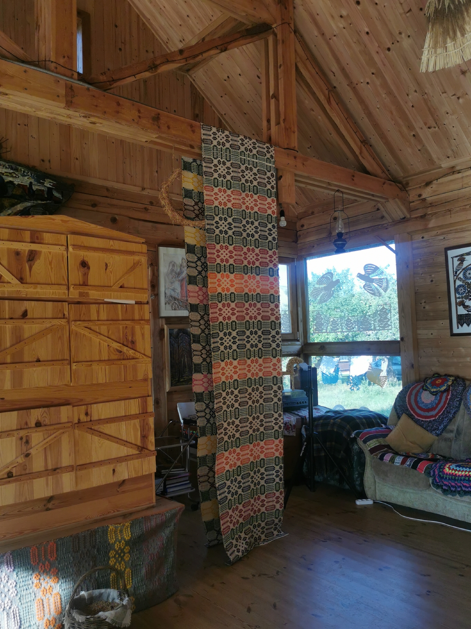 The image shows the interior of a rustic wooden cabin with a sloped ceiling and exposed wooden beams. A long, woven textile with a geometric pattern in red, black, and white hangs from the ceiling in the center of the frame. To the left, a large wooden cabinet with multiple drawers is visible. In the foreground, a woven tapestry covers the front of a wooden bench, with a basket containing grain placed on top. To the right, a sofa is draped with colorful crocheted blankets and cushions. A large window with a view of trees and grass is behind the sofa, adorned with cut-out bird and tree silhouettes. The flooring is polished wood. The scene depicts a cozy, handmade interior. The location is specified as Шчарчоўскі сельскі Савет, Belarus.