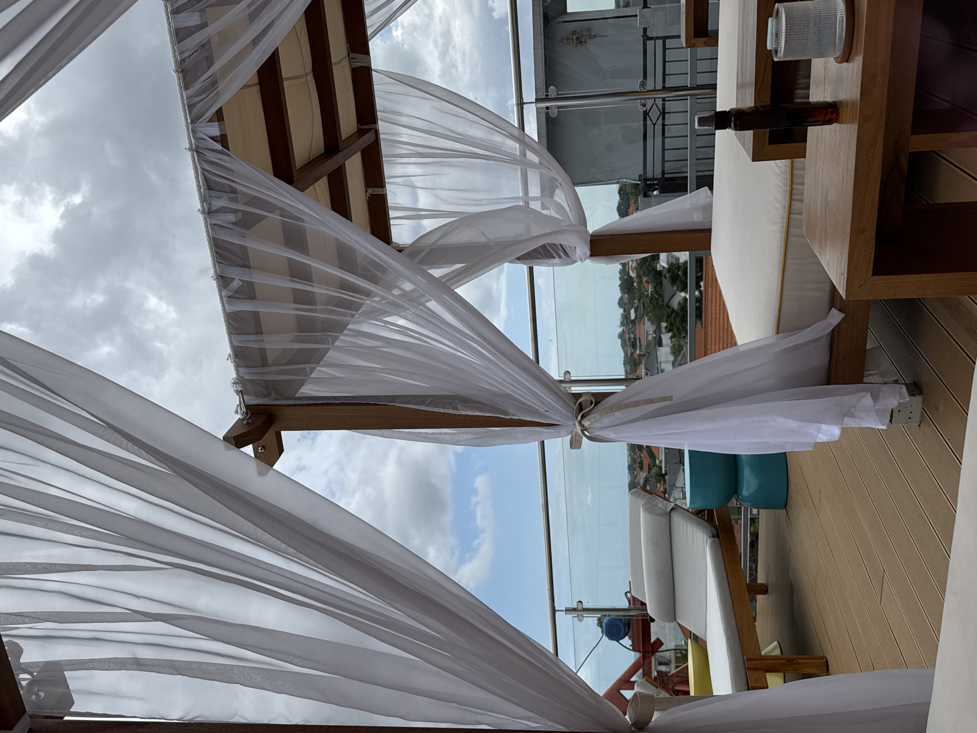 The image shows a rooftop lounge area in North Kuta, Indonesia. The scene is dominated by white sheer curtains draped around a wooden structure, creating a canopy. In the foreground, a wooden table holds a glass of water and a dark glass bottle with a spray nozzle. To the left, a white cushioned chaise lounge is positioned on a wooden deck. Behind the chaise lounge, a bright teal cylindrical stool is visible, along with a railing and a glimpse of residential buildings in the distance. The sky is overcast with patches of blue sky and clouds. The overall atmosphere suggests a tranquil outdoor relaxation space.