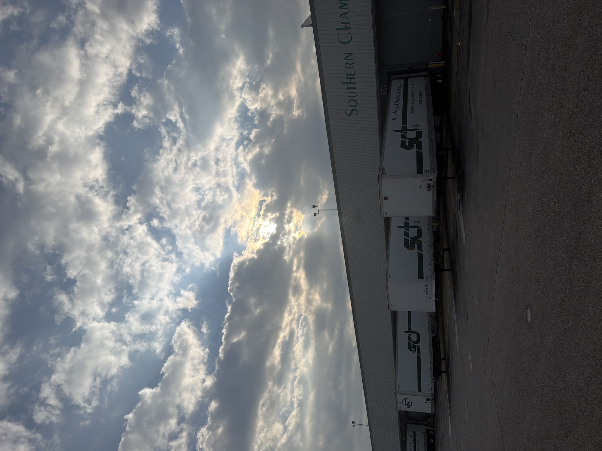 The image depicts the exterior of a commercial building, likely a warehouse or distribution center, in Chattanooga, Tennessee, United States. The building has a long, grey, corrugated metal facade. The name "SOUTHERN CHAMPION" is visible in large green lettering across the top of the building. Parked adjacent to the building are several white semi-trailer trucks, some bearing the logo "SCI" in green. The trailers are positioned at loading docks, indicated by the raised platforms and doors. The ground in front of the building is paved asphalt. Above the building, the sky is filled with dramatic clouds, with sunlight breaking through in several areas, creating a partly cloudy and somewhat dramatic sky. The overall scene suggests a working industrial or commercial environment under a dynamic sky.