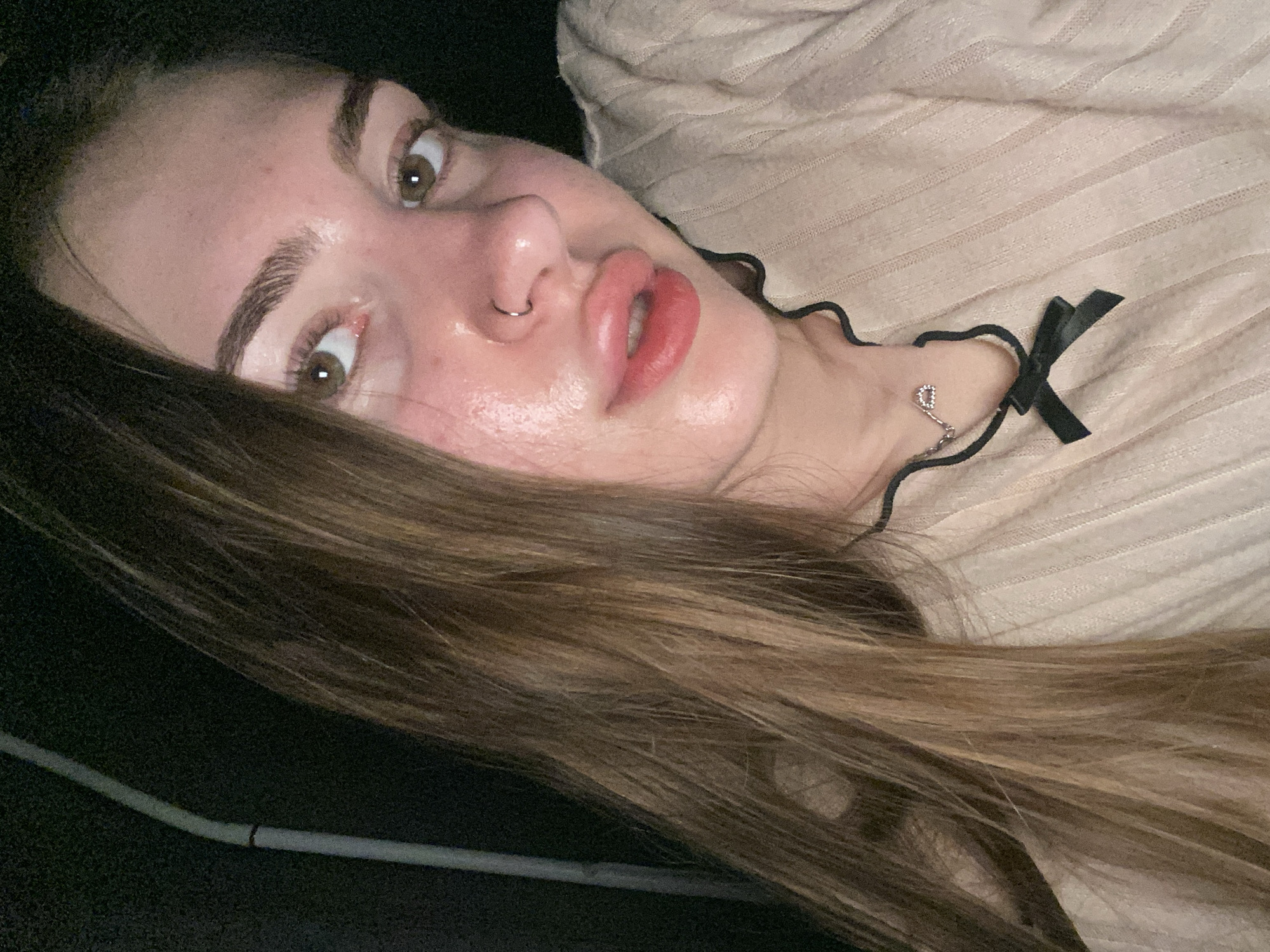 A young woman with long brown hair and light skin is taking a selfie in a dimly lit outdoor environment. She is wearing a beige, ribbed sweater with a dark trim and a small bow at the neckline. She has a nose ring and a delicate necklace with a small pendant. Her lips are slightly parted and appear to be glossed. The background is dark, with a vertical object that appears to be a light pole to the left of the frame. The overall lighting suggests it is nighttime. The image does not contain any information that can indicate the specific location within Дьяково, Russia.
