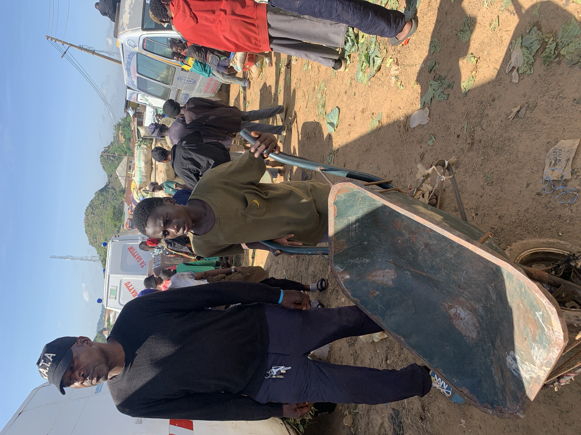 An outdoor scene depicts a group of people in what appears to be a marketplace or roadside gathering in Gura Top, Nigeria. The ground is dusty and scattered with green leafy vegetables and some plastic debris. In the foreground, a young person wearing a green t-shirt is pushing a green wheelbarrow. To the left, an adult male wearing a black long-sleeved shirt and a black cap with "G.A.T.A." is standing, looking off to the left. Several other individuals, including children and adults, are visible in the background, some near white vans. One van has text in what appears to be Arabic script. The sky is clear and blue, with a few hills and buildings visible in the distance. A telecommunications tower is also present in the background. The overall impression is one of a busy, somewhat disorganized community setting.