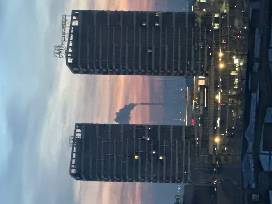 The image displays an urban twilight scene in Krasnoyarsk, Russia. Two prominent, dark, multi-story rectangular high-rise buildings stand centrally. The building on the right features an illuminated "METROPOLIS" sign and a distinctive logo on its roof. The left building has a similar structural element on its roof. Scattered illuminated windows are visible on both towers.
Between and behind these two structures, a tall, slender industrial chimney is visible in the mid-distance, emitting a substantial plume of white smoke. This chimney is identifiable as belonging to the Krasnoyarsk Thermal Power Plant-2 (TPP-2). The sky above exhibits a gradient, transitioning from light blue at the zenith to shades of pink and orange near the horizon.
Below the main towers, the cityscape unfolds with numerous artificial lights, indicating active commercial areas and thoroughfares. Partially visible signage includes a red sign with "ome" discernible. The foreground exhibits visual distortions, including vertical wavy lines and reflections, consistent with the image being captured through a pane of glass. No individuals are depicted.