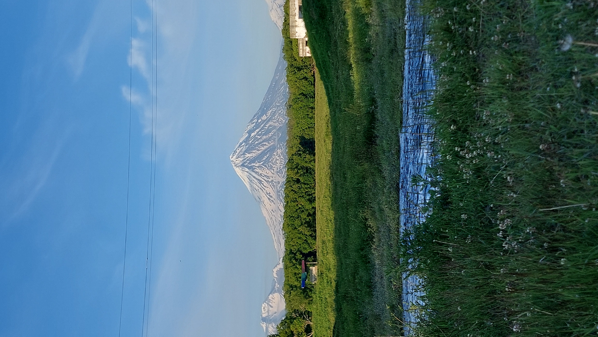 A picturesque natural landscape unfolds with a snow-capped stratovolcano dominating the background. The volcano, with its distinctive conical shape, is partially covered in snow, contrasting with the clear blue sky above, which is streaked with wispy white clouds. In front of the volcano, a dense line of green trees forms a verdant backdrop, suggesting a lush, forested area.
Below the tree line, a grassy meadow stretches towards the foreground, illuminated by the warm glow of sunlight. A small, open-sided structure with a colorful roof is visible within the meadow, possibly a shelter or pavilion. To the right of the meadow, a weathered, abandoned-looking building with concrete walls stands on an elevated incline, hinting at past human activity.
In the immediate foreground, a body of water, likely a river or stream, flows across the frame. The water's surface is rippled, reflecting the light and adding a dynamic element to the scene. The banks of the waterway are covered in tall green grass, interspersed with small white wildflowers, creating a textured and natural foreground. The scene is devoid of any human figures. The overall impression is one of serene natural beauty, with the majestic volcano as the focal point.
