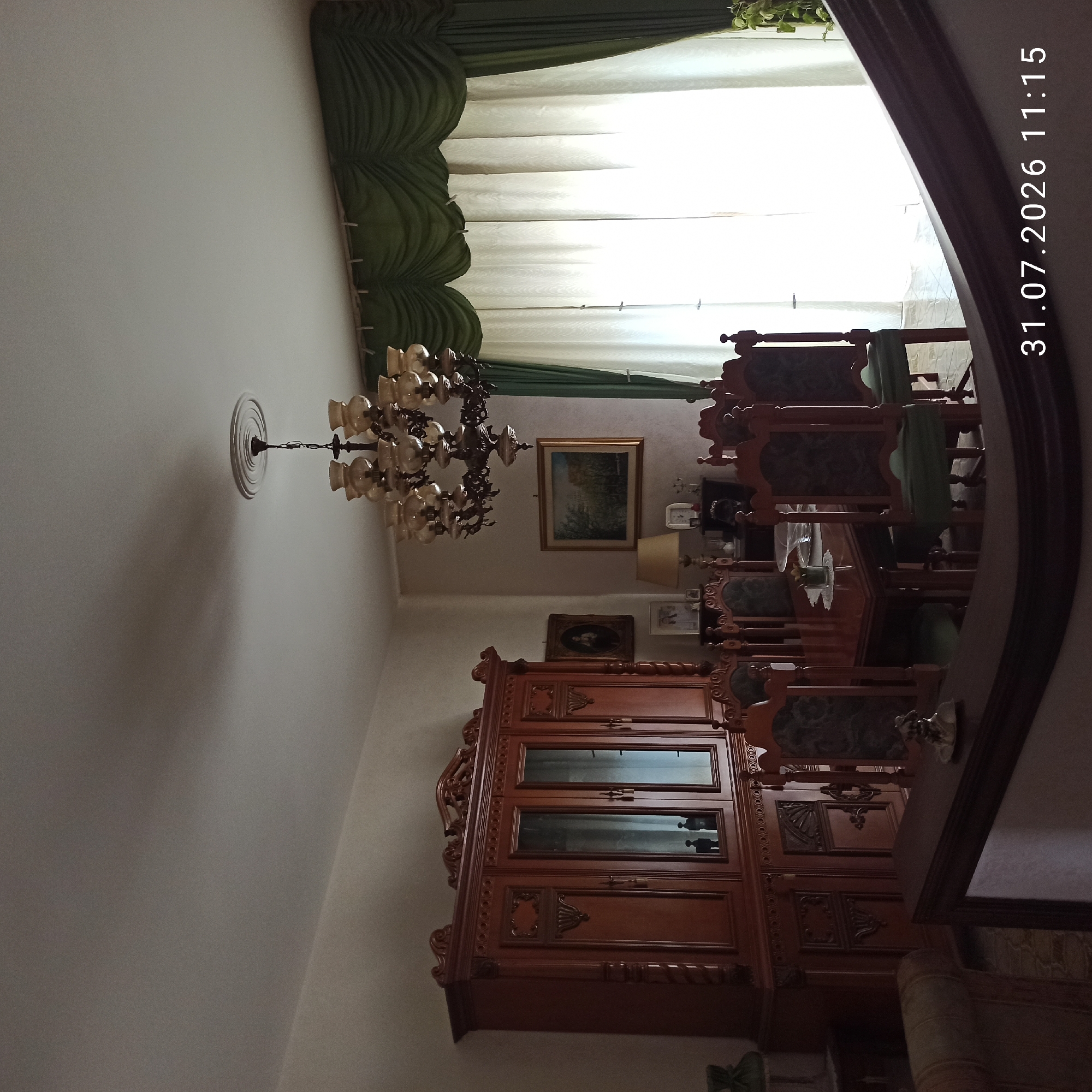 The image depicts a formal dining room in Modugno, Italy, characterized by ornate wooden furniture. A large, dark wood china cabinet with glass doors stands prominently on the left side of the frame. In the center, a dining table is set with chairs, all featuring dark wood frames and upholstered seats. A chandelier with multiple glass shades hangs from the ceiling, and green draped curtains frame a brightly lit window on the right. The overall impression is one of a traditional and well-maintained dining space. The timestamp "31.07.2026 11:15" is superimposed on the bottom right corner of the image.