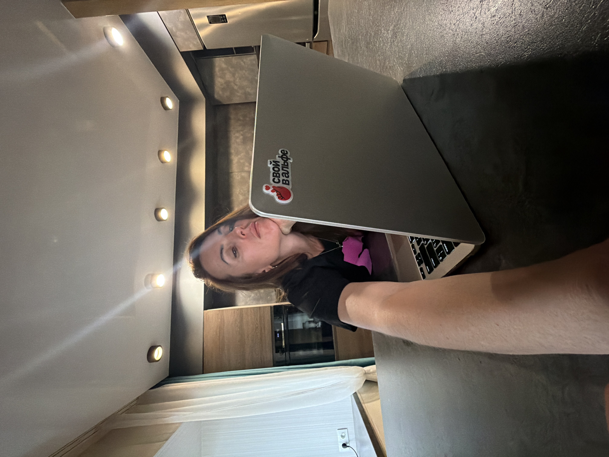 An interior photograph, rotated 90 degrees clockwise from its natural orientation, shows a woman partially visible at a dark-colored countertop, likely in a modern kitchen or living space in Birobidzhan, Russia. The woman, with brown hair and a neutral to slightly pleasant expression, looks directly at the camera. She is wearing a black top with a pink design visible on her left side and a necklace. Her right arm extends onto the dark countertop, and her upper body is positioned near an open silver laptop.
The laptop, placed on the dark surface, has a sticker on its closed lid that reads "СВОЙ ВАЛЬФЕ" (SVOY VALFE) in white letters on a black background, accompanied by a red heart-shaped icon, possibly representing a drop of liquid. The keyboard is partially visible beneath her arm.
The background reveals modern interior details. Above the woman, several circular, recessed or surface-mounted spotlights illuminate a light-colored ceiling or cabinet underside, casting warm yellow light. Further back, wooden cabinets with a sleek, integrated oven or microwave are visible, hinting at a kitchen area. Below this, a light blue or teal curtain and what appears to be a white piece of furniture (possibly a bed or sofa) and a white wall outlet with a black cord are present. The room appears to be lit by artificial light, suggesting it is evening or night. The overall aesthetic is contemporary and functional.
