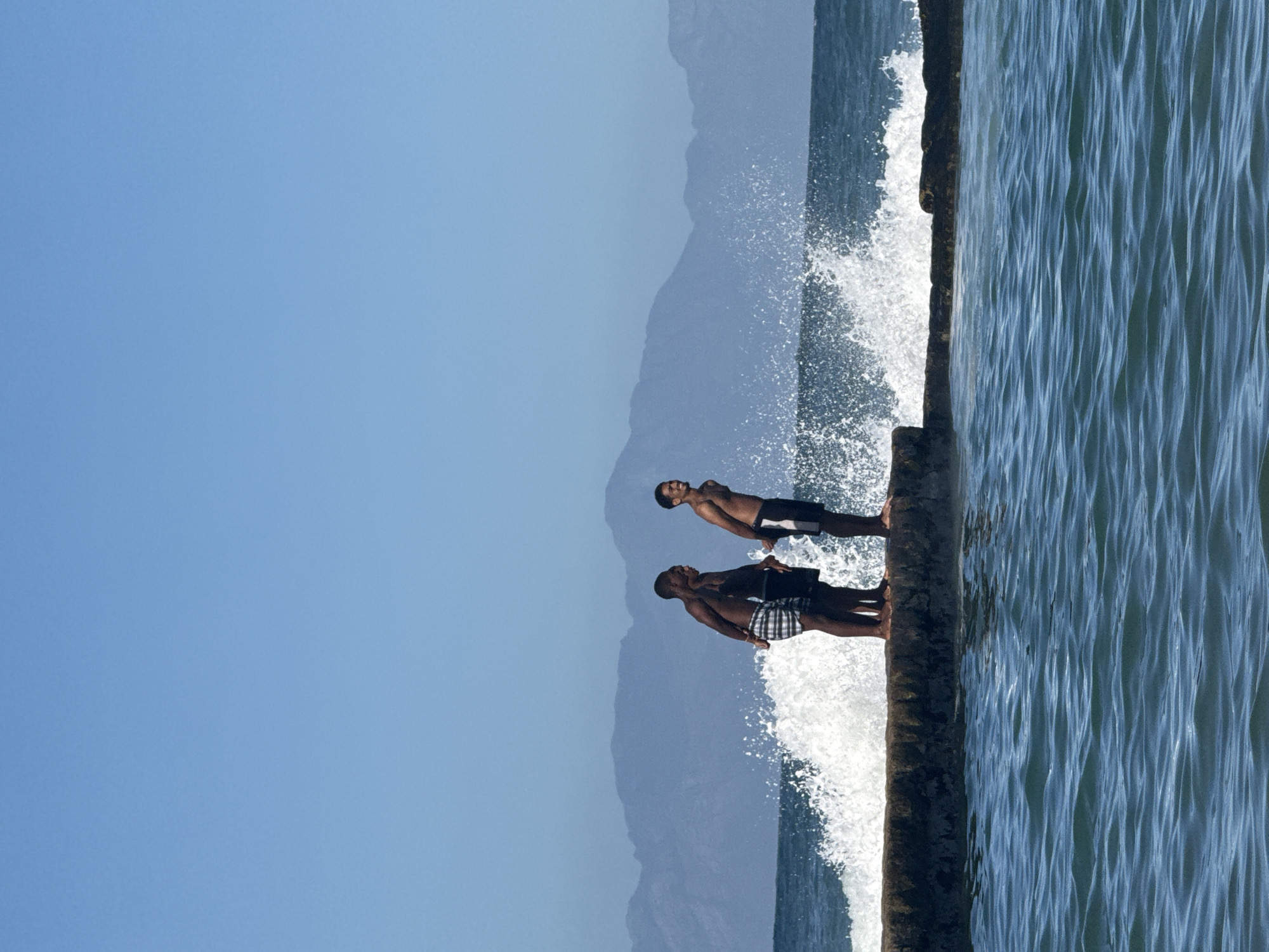 Two men stand on a concrete pier as waves crash against it. The sky is a clear blue, and mountains are visible in the background. The location is identified as Cape Town, South Africa.