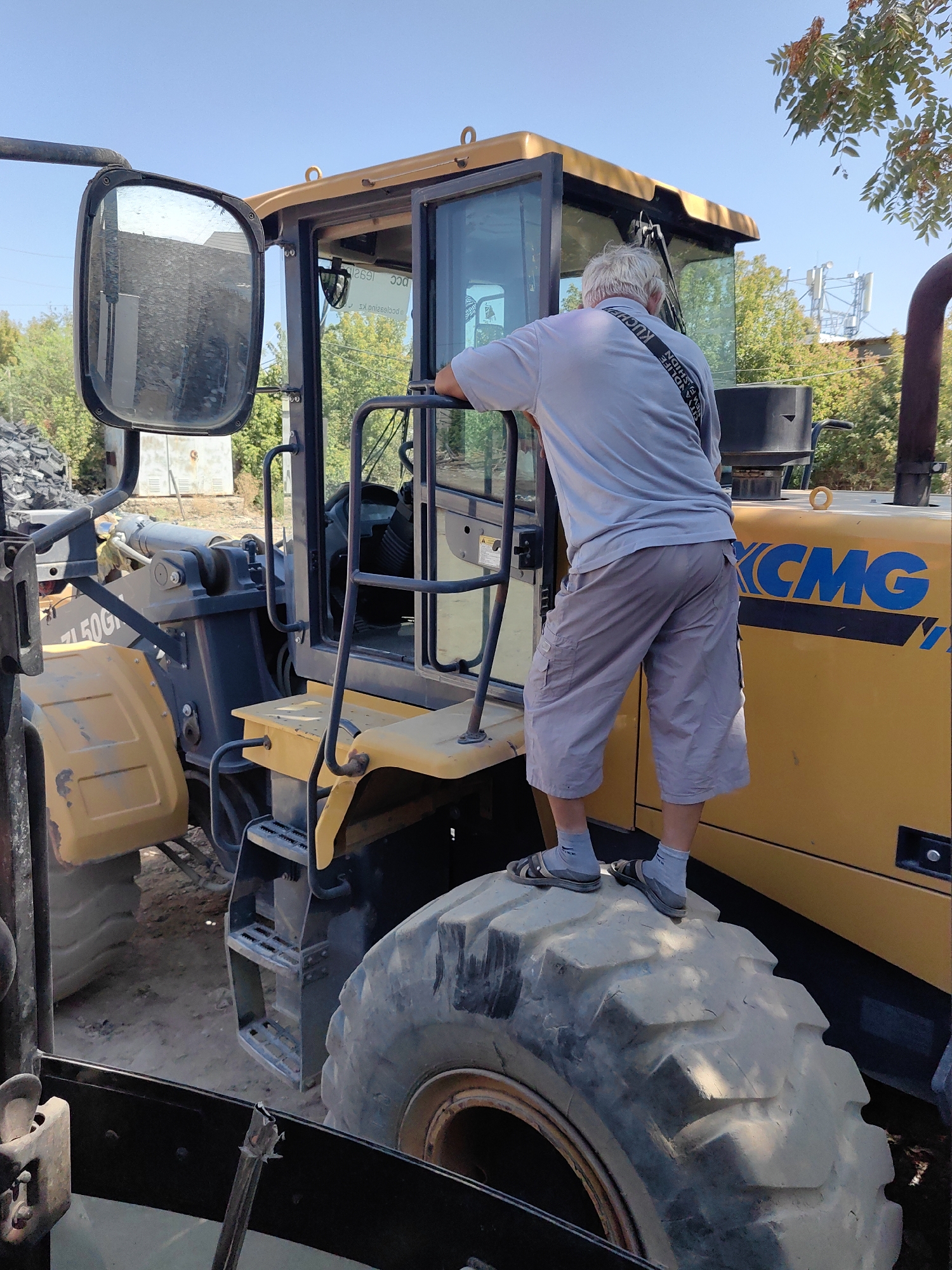 A man with gray hair stands on the large tire of a yellow construction vehicle, leaning his arms on the cab. He is wearing a light gray long-sleeved shirt and light gray cargo shorts, along with dark sandals and light gray socks. The construction vehicle appears to be a loader, with "CMG" visible in blue lettering on its side. The scene is outdoors, with trees and buildings in the background. The ground is dirt and gravel. The sky is clear and blue. The image was taken in Shymkent, Kazakhstan, likely at a construction site or depot.