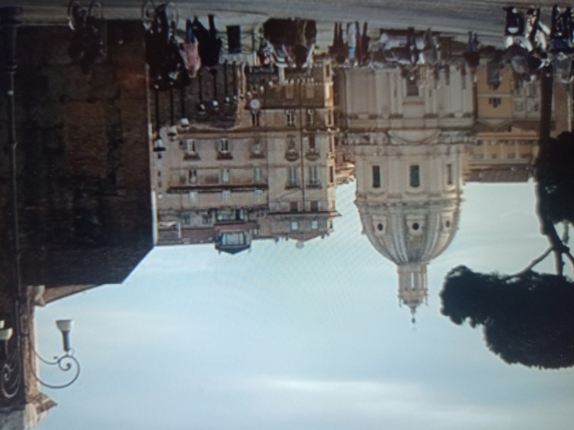 This is a blurry, pixelated image depicting a street scene with a large, domed building that resembles a church or cathedral on the left. In the background, there are several multi-story buildings with many windows, suggesting an urban environment. In the foreground, there are several people walking, some on a street where motorcycles and a car are visible. A decorative street lamp with a distinctive curved arm is present on the right side of the image. The sky is light blue with some hazy clouds. The overall impression is of a sunny day in a European city. The image is not a city in Mukachevo, Ukraine.