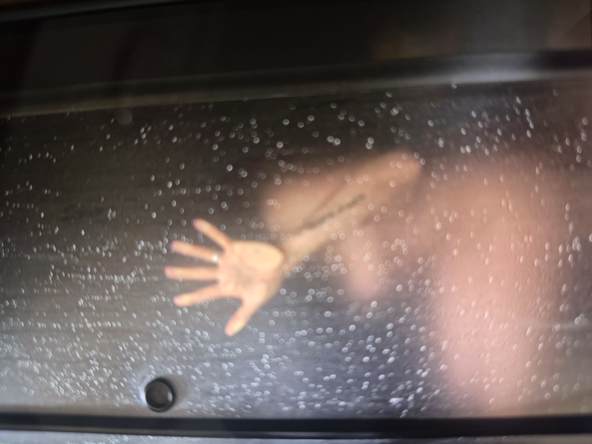 The image shows a blurred, seemingly nude figure behind a wet glass surface. A hand, with a ring on one finger, is pressed against the glass. The background behind the figure is dark, and the glass has many water droplets obscuring the view. A black frame surrounds the glass. A drain is visible at the bottom. The location is Unknown, Unknown.