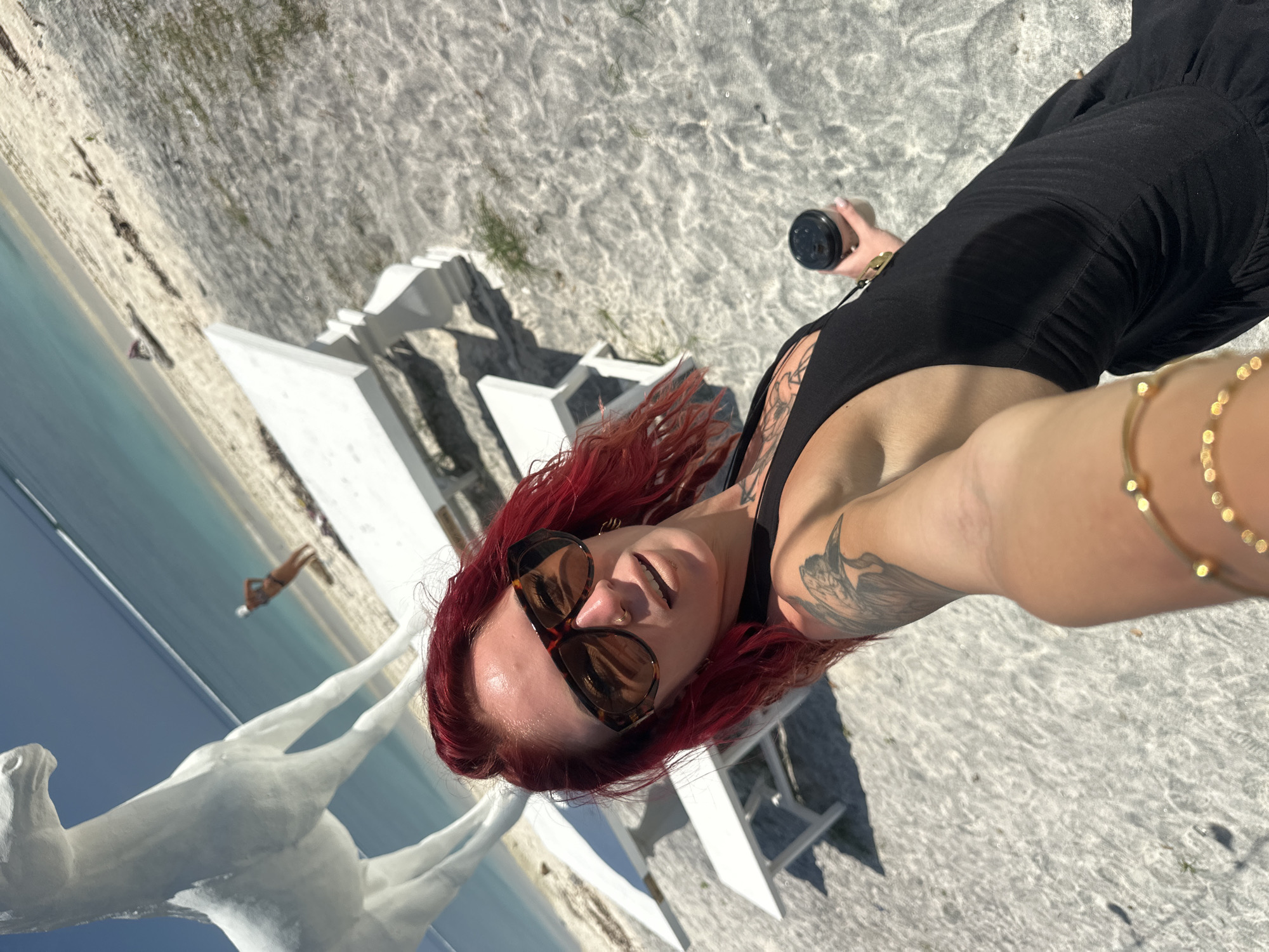 The image depicts a person with red hair and sunglasses, wearing a black tank top and a gold bracelet, taking a selfie. The person is holding a black container in their left hand. They have tattoos on their left arm and chest. The background shows a beach with white sand and turquoise water. There is a white sculpture of two horses and white furniture in the scene. A person is visible in the distance on the beach. The location is the city of Unknown, Indonesia.
