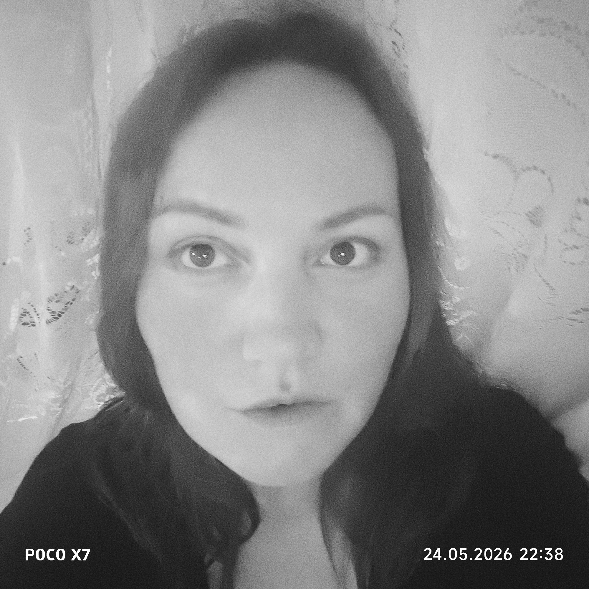 A grayscale close-up photograph depicts a person with long, dark hair. The person is facing directly towards the camera, with their eyes open. Behind the person is a patterned material, potentially a curtain or wall covering. The bottom left corner of the image reads "POCO X7" and the bottom right corner reads "24.05.2026 22:38". The location is Unknown, Unknown.