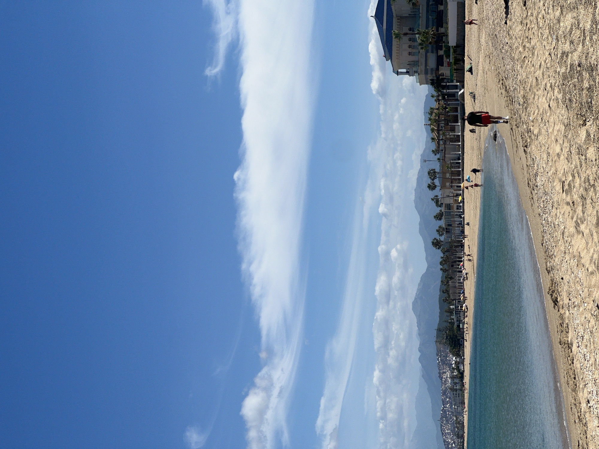 This image shows a beach in M'Diq, Morocco, with the sea and sky dominating the view. The sky is clear with some wispy clouds. Along the beach, there are several buildings, including a multi-story structure. People are walking along the shoreline. The sand is light-colored, and the water appears calm and clear. Mountains are visible in the background, behind the buildings.