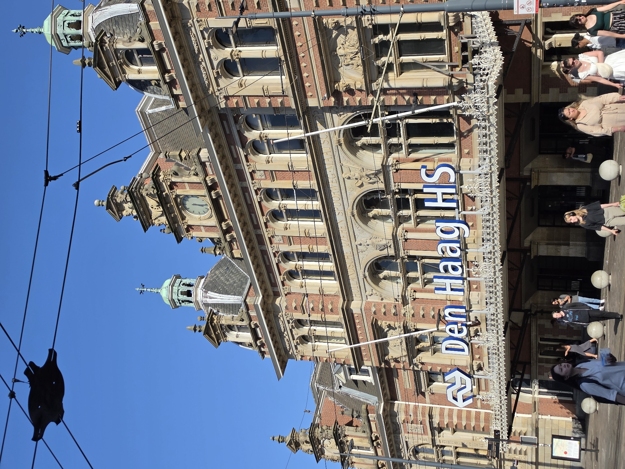 The image shows the exterior of Den Haag HS station in The Hague, Netherlands. The grand red-brick building features ornate architectural details, including arched windows, decorative carvings, and cupolas. A large sign above the entrance reads "Den Haag HS" in blue letters. Several people are walking on the sidewalk in front of the station, some are looking at the camera, and others are walking past. The sky is clear and blue, and the sun is shining, casting shadows on the building and the street. There are overhead wires and a dark, star-shaped object suspended from them in the upper left portion of the image, likely part of the overhead electrical system for trams or trains.