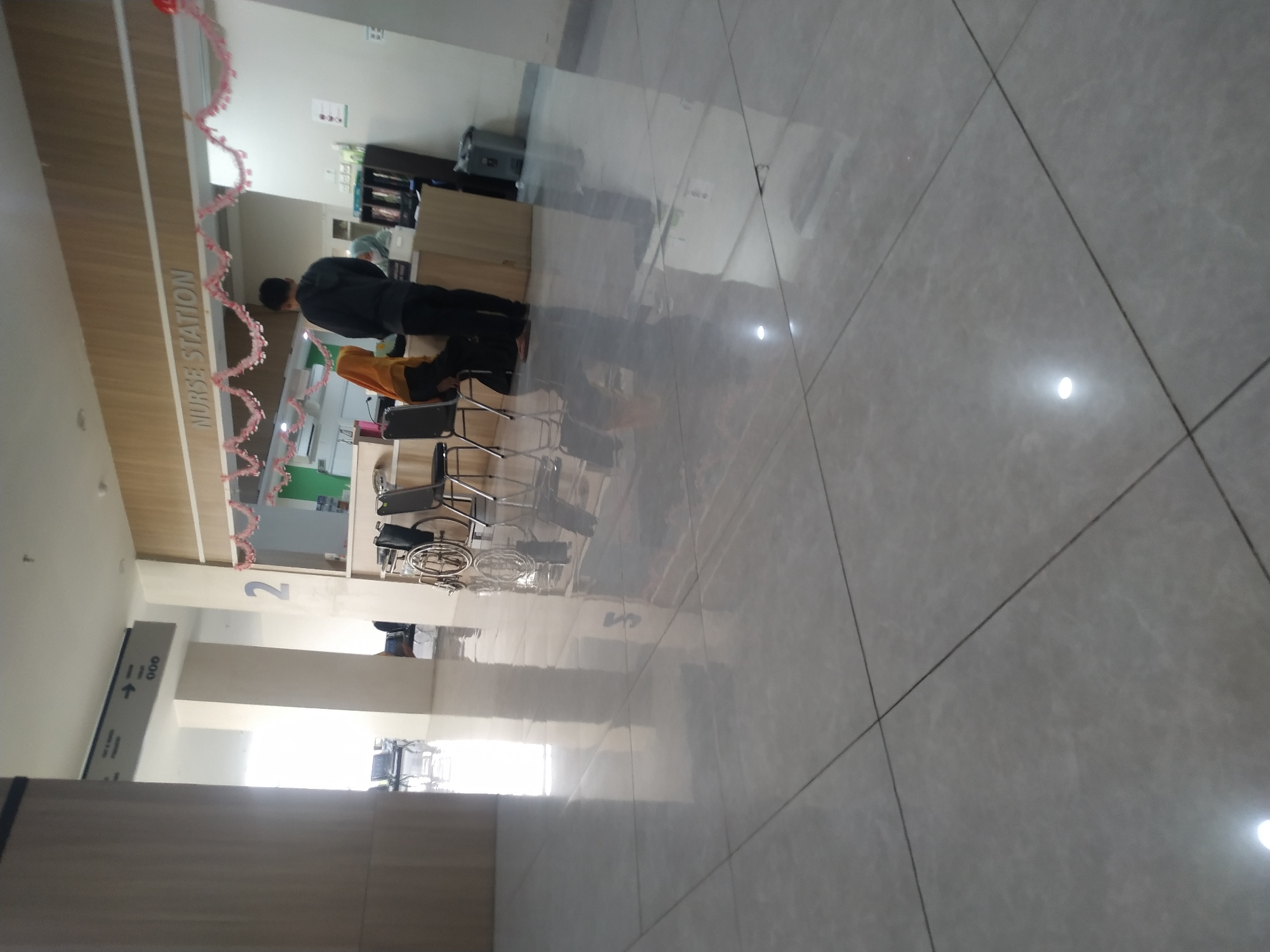 The image depicts the interior of a hospital or clinic, likely in Mojosongo, Indonesia, specifically at a nurse's station. The environment is clean and modern, with polished, reflective tile flooring that shows significant glare from overhead lighting. A directional sign with the number "000" and an arrow pointing right is visible on a column. To the right, a "NURSE STATION" sign is mounted above a counter. Two individuals are standing at the counter, one wearing a yellow hijab and the other a dark hoodie. There are several chairs and a wheelchair present in the vicinity. The overall scene suggests a functional healthcare setting with ongoing activity.