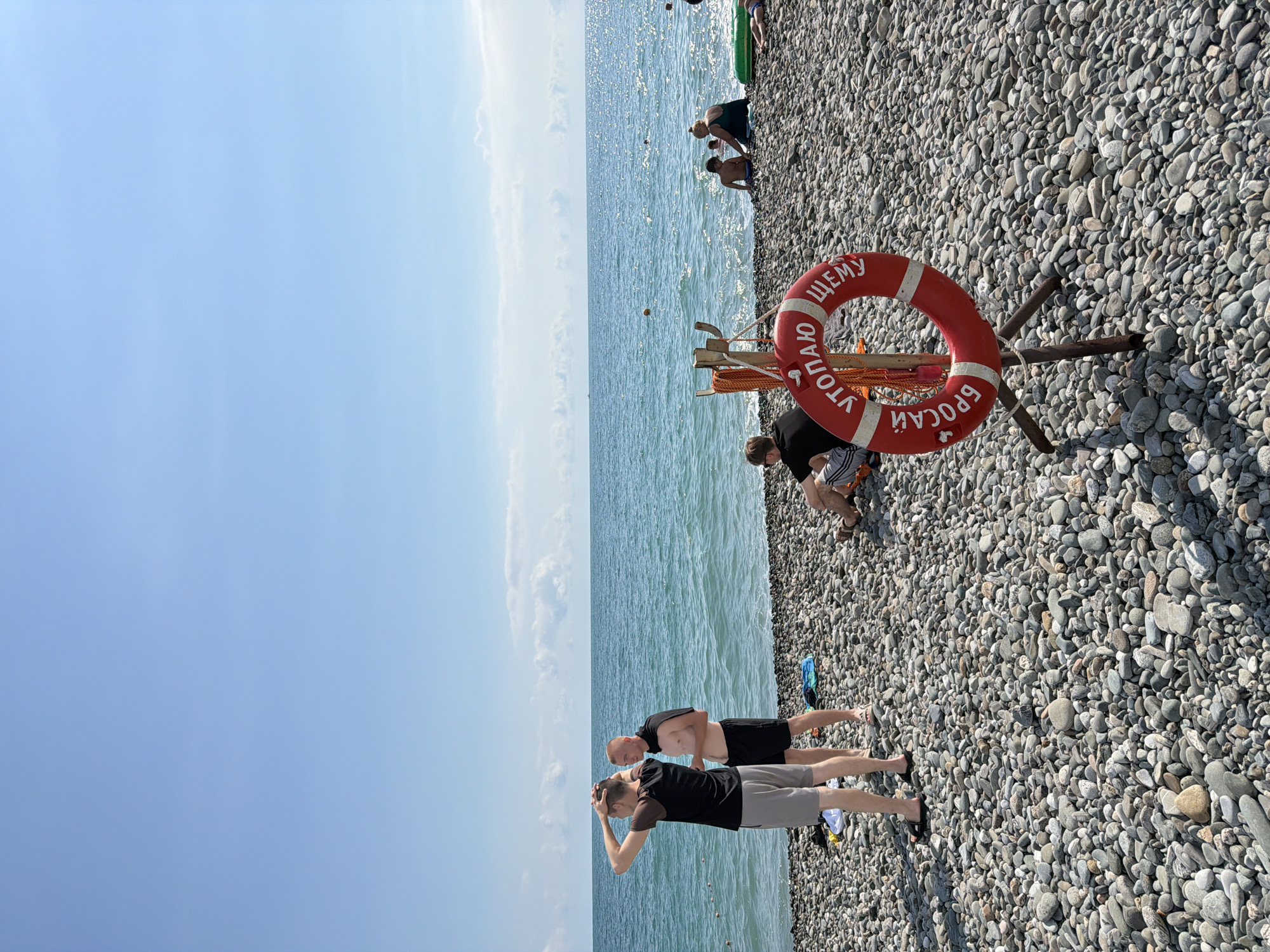 The image depicts a sunny day at a pebble beach in Sirius, Russia, adjacent to the Black Sea. The water is a vibrant turquoise, with gentle waves and visible sunlight glinting off the surface. Several people are scattered across the beach and in the shallow water. In the foreground, a red life preserver with white stripes is mounted on a wooden stand. Russian text, "УТОПАЮ ЩЕМУ БРОСАЙ" (meaning "help, drowning, throw!"), is visible on the life preserver.
Two men stand near the water's edge, facing away from the camera, with one man touching his head. Further back, a group of people, including children, are seated or playing near the shoreline. Another individual is seated on the beach in the foreground, facing away from the camera, wearing sunglasses. The beach is covered in smooth, grey pebbles of various sizes. The sky is a clear, bright blue with a few wispy clouds on the horizon.