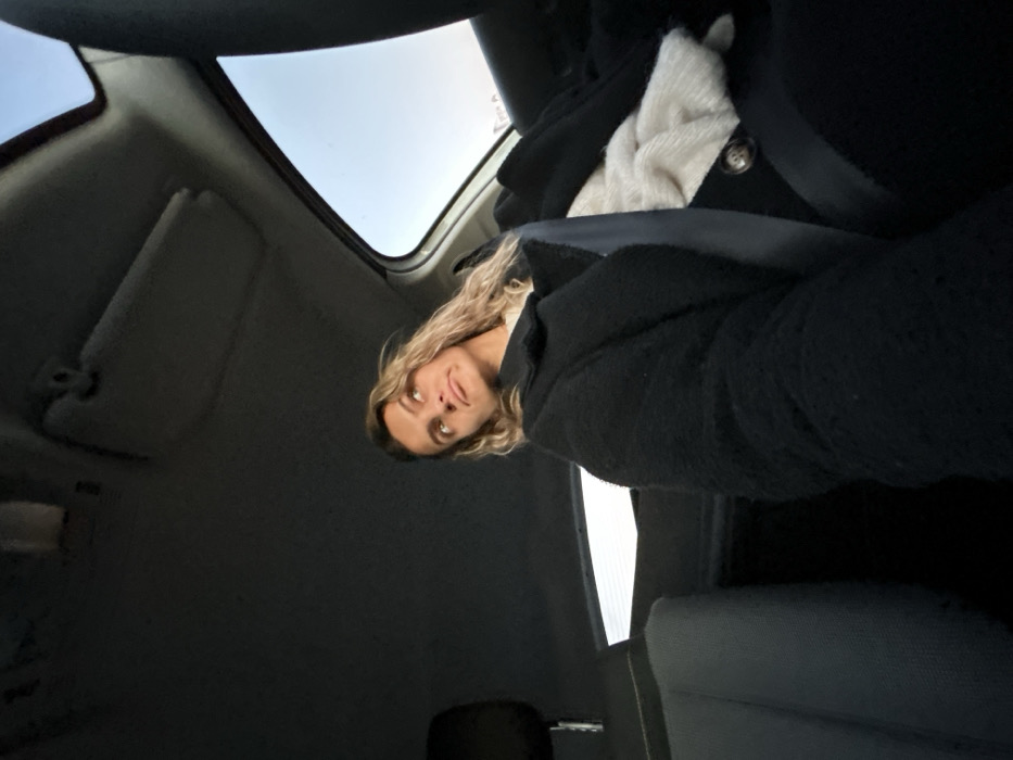 A person with long blond hair sits in a car, looking up toward the camera. They wear a black coat over a white shirt or sweater. A seatbelt crosses their chest. The interior of the car is dark, with the sunroof visible in the top left corner. The car's sun visor is also visible. The camera angle is from below, looking up toward the subject.