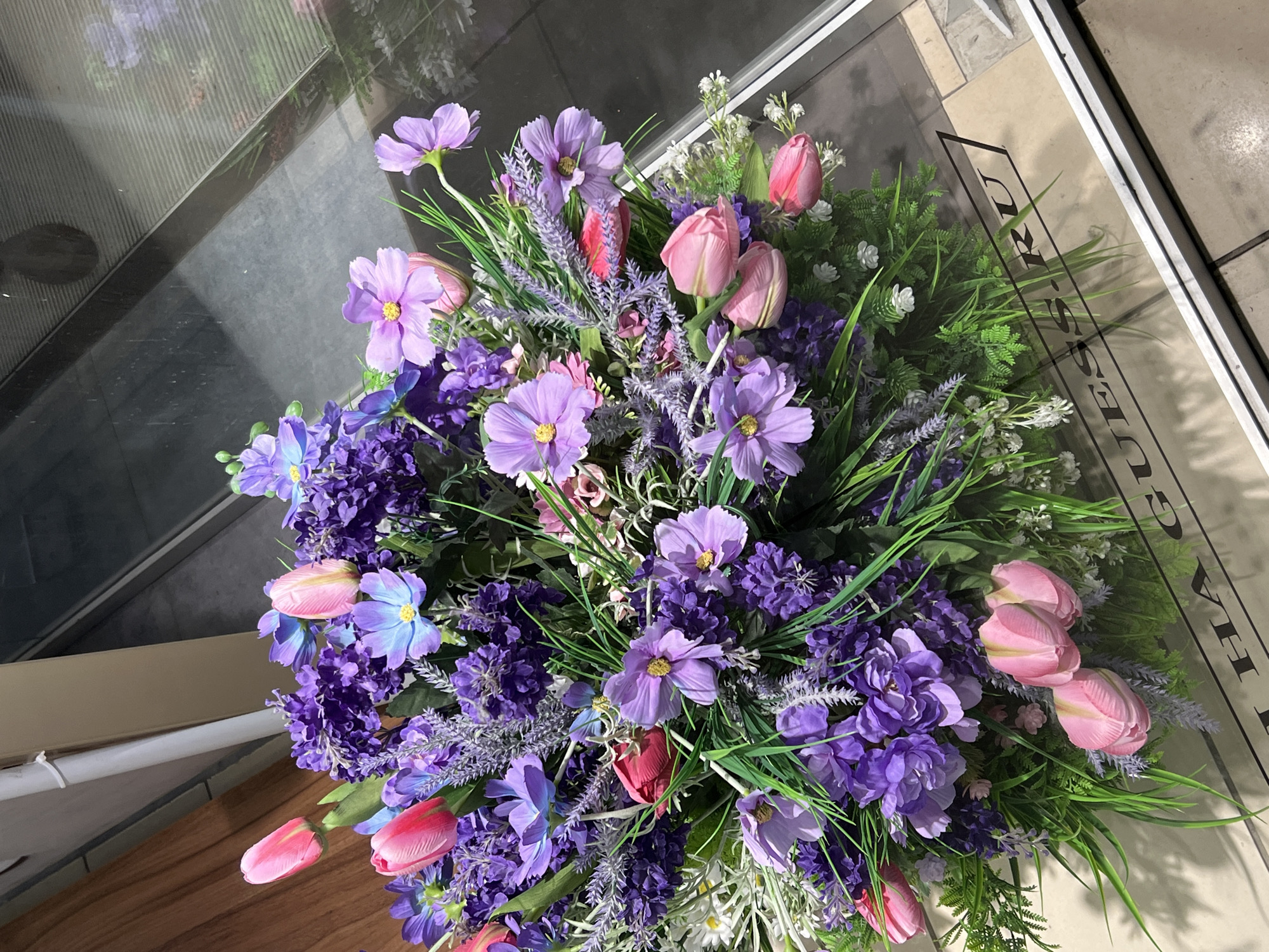 A bright, overhead shot displays a lush artificial flower arrangement, predominantly featuring shades of purple, pink, and blue, with green foliage. The arrangement is positioned within a white-bordered rectangular frame, likely a display case or a planter. The background shows a wooden surface on the left and a reflective surface, possibly glass, on the right, which appears to be a shop window or door. Through the glass, a blurred urban environment is visible, suggesting the arrangement is located in a commercial setting. The text "I HA GUESS.RU" is visible on a light-colored surface beneath the arrangement, suggesting it is a display for a retail store or a brand, potentially Guess. While the image was taken in Kazan, Russia, no specific landmarks are identifiable. There are no people or animals present in the scene.