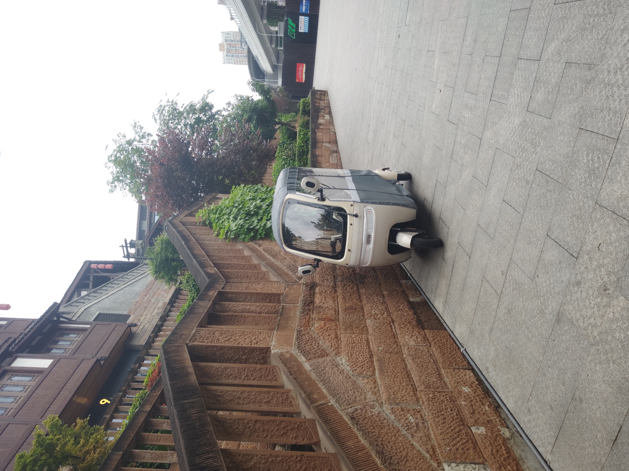 The image shows a three-wheeled vehicle parked on a sidewalk in Shapingba District, China. The sidewalk is made of light gray paving stones. Adjacent to the sidewalk is a stone wall with a staircase leading upwards. Vegetation is visible behind the wall and along the staircase. A traditional Chinese building is partially visible to the left of the staircase, and modern buildings are in the background across the street. The sky is overcast. No people are visible in the image.