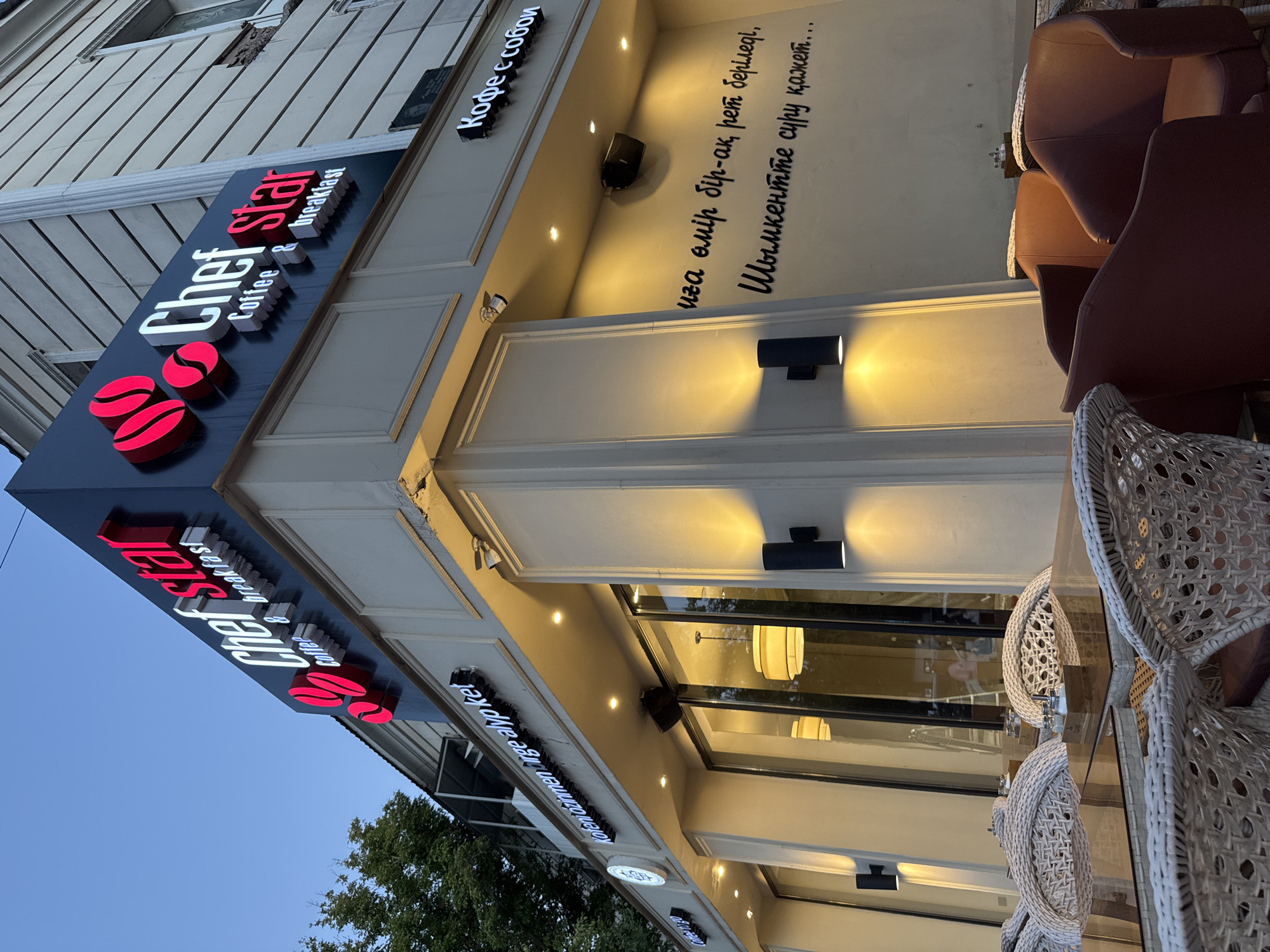 The image shows the exterior of a cafe named "Chef Star Coffee & Breakfast" located in Shymkent, Kazakhstan. The cafe has a prominent illuminated sign featuring the cafe's name in red and white lettering, along with red coffee bean graphics. Below the main sign, there are other signs with text in what appears to be Kazakh and Russian, such as "Кофе с собой" (Coffee to go) and a longer inscription on the building's facade in a script that may be Kazakh.
The facade of the building is cream-colored with decorative molding. There are several outdoor seating areas with tables and chairs. Some chairs are made of white wicker, while others are upholstered in a reddish-brown material. The tables have glass tops and appear to be set with salt and pepper shakers. Two black wall-mounted lights illuminate the facade. The reflection in the cafe's windows shows the interior, with pendant lights and what looks like a ladder and some people working inside. The sky in the background is a dusky blue, suggesting it is either early morning or late evening. Trees are visible on the left side of the image.