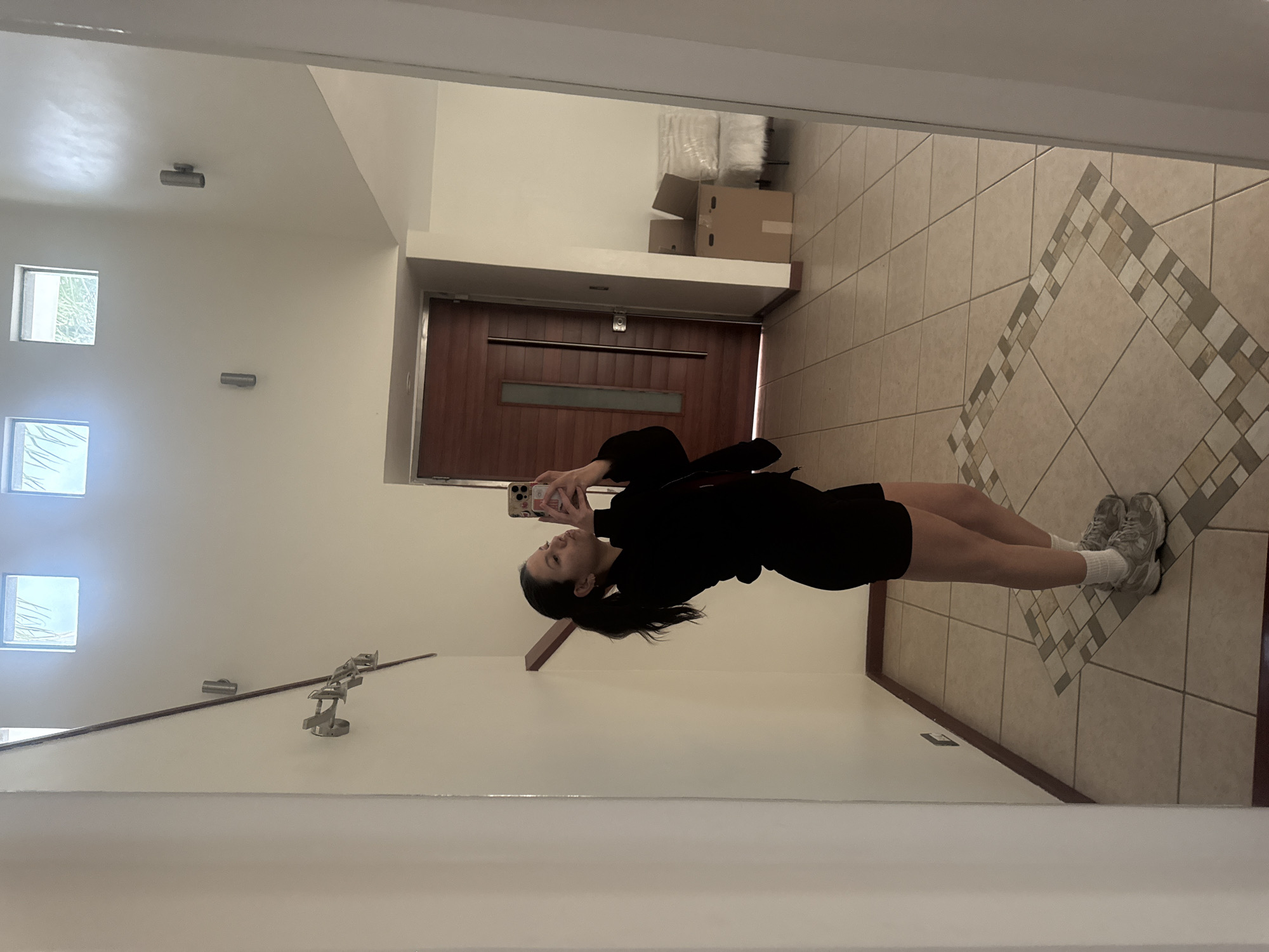 A young woman is taking a selfie in a mirror inside a home, presumably in Heroica Puebla de Zaragoza, Mexico. She is wearing a black top and shorts, white socks, and gray sneakers. The background features a tiled floor with a decorative pattern, a dark wood door with a window, and boxes stacked on a ledge. The walls are white with visible lighting fixtures and windows. The mirror reflects the interior of the home.