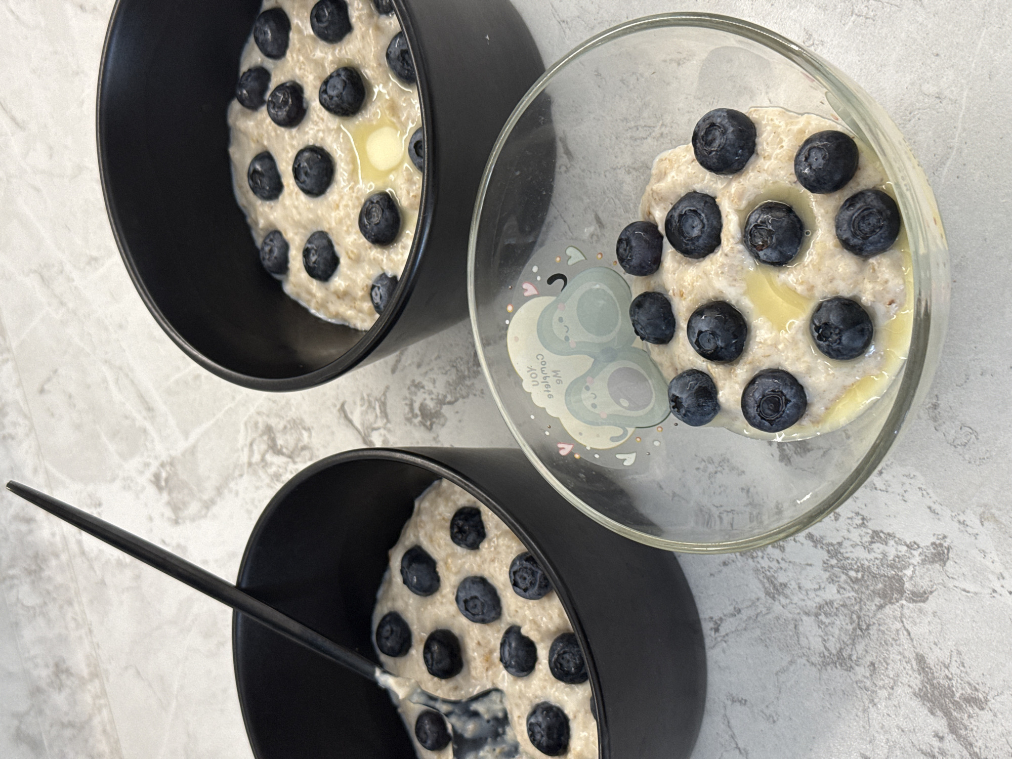 The image shows three bowls of oatmeal with blueberries and melted butter, arranged on a marbled countertop. Two black bowls are positioned to the left and right, with a clear glass bowl in the foreground. A black utensil rests in the left bowl, indicating it has been partially consumed. The countertops and background are consistent with an indoor setting, likely a kitchen. There are no people visible in the image. The scene does not contain any information to place it in Tomilino, Russia.