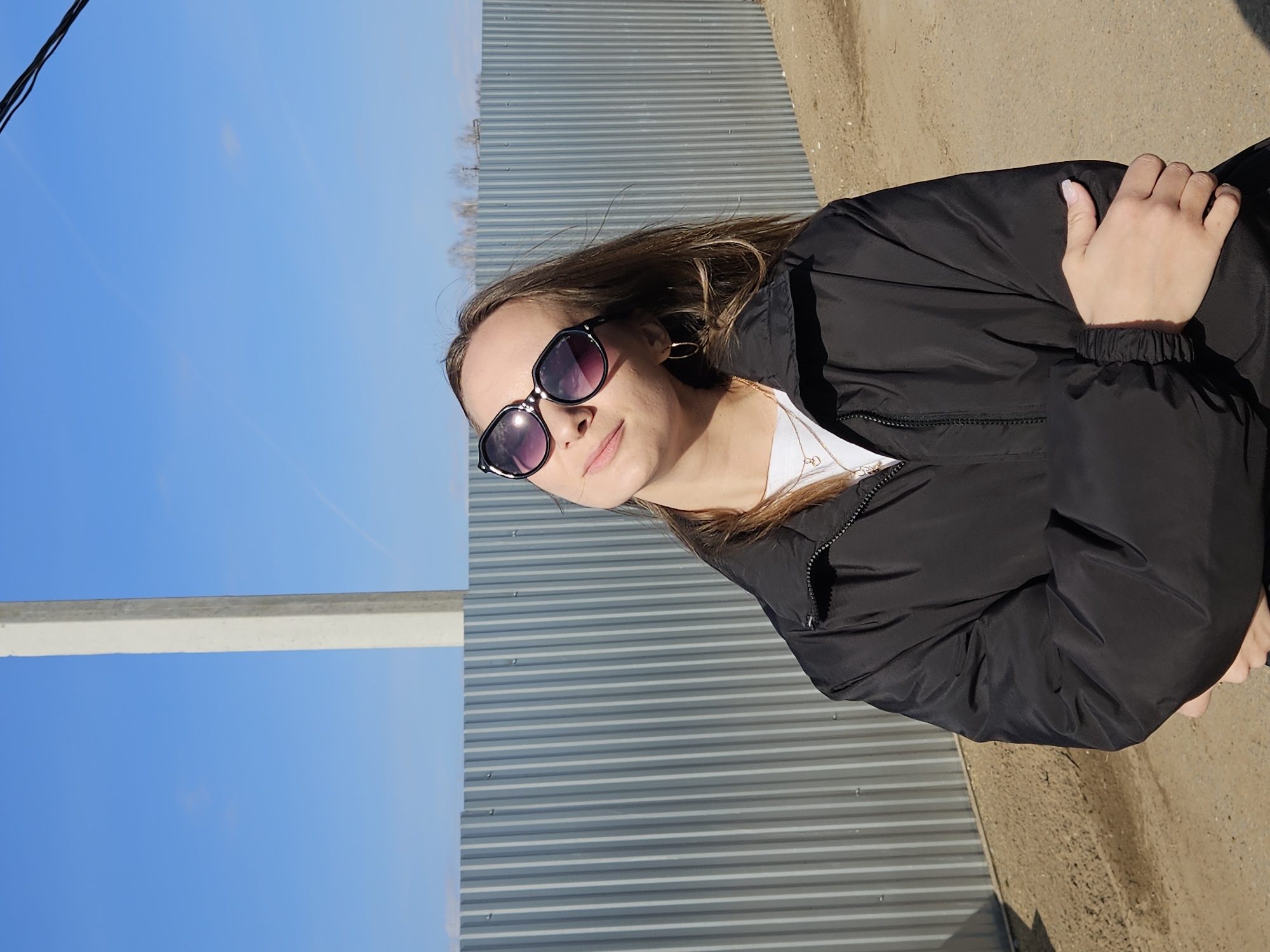 A young woman is captured posing outdoors on a bright, sunny day in Chaikovsky, Russia. She is depicted from the chest up, with her head slightly tilted and a calm, pleasant expression, looking directly at the viewer. Her long, light brown hair frames her face, and she is wearing dark, black-framed sunglasses. She is dressed in a black lightweight jacket over a white top, accessorized with a delicate necklace and hoop earrings. Her left hand, with light-colored manicured nails, rests gently on her right arm.
The background features a prominent grey corrugated metal fence or wall with horizontal ridges, hinting at an industrial or utilitarian outdoor setting. A clear, vibrant blue sky occupies a significant portion of the scene to her left. A white concrete beam or pillar is visible on the far left edge of the frame. To her right, the ground appears to be a light brown, sandy, or dirt patch. In the distance, just above the corrugated fence, faint outlines of bare trees or bushes suggest a cooler season, such as late autumn or early spring. The scene is well-lit by natural sunlight, indicating it was taken during the day. There is no visible text in the image.