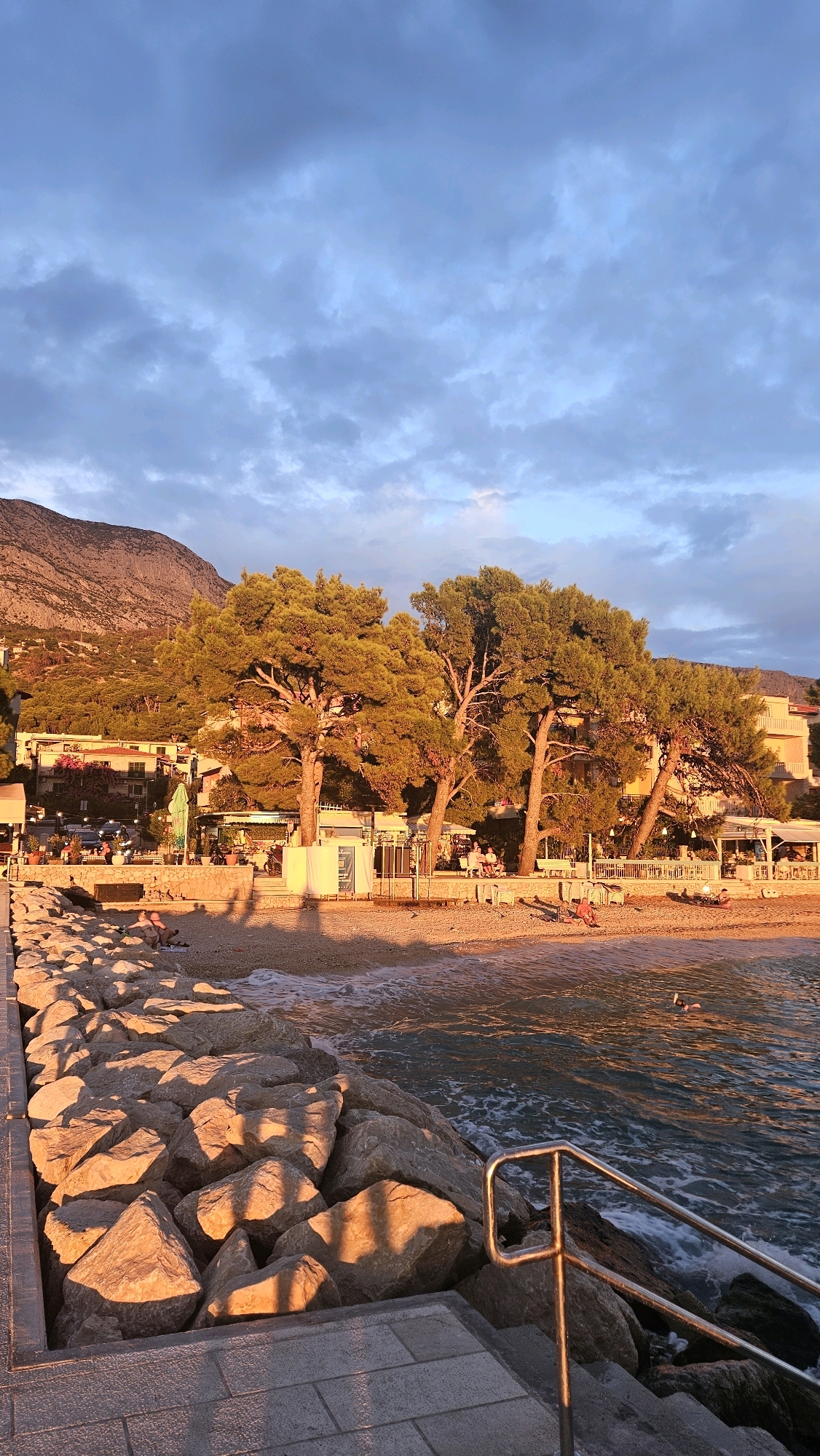 The image depicts a coastal scene at sunset in Općina Tučepi, Croatia. A rocky breakwater with paved steps and a metal railing leads towards the sea. The beach is lined with several large pine trees casting long shadows. Beyond the trees, there are buildings, possibly hotels or apartments, with awnings and outdoor seating areas. People are visible on the beach, some sunbathing on the sand, and one person is swimming in the sea. The sky is partly cloudy with warm light from the setting sun illuminating the scene, casting a golden glow on the trees, beach, and rocks. The sea is calm with gentle waves lapping the shore.