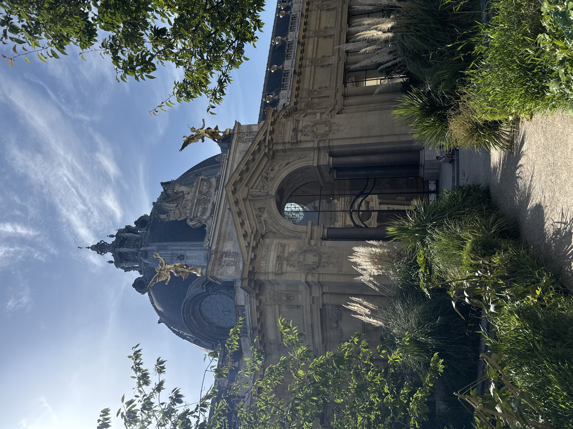 The image captures the Petit Palais in Paris, France, on a clear day with a few wispy clouds. The grand Beaux-Arts building is adorned with a prominent domed roof and classical architectural details. Two golden winged statues are perched atop the building's facade, overlooking the scene. Lush green trees and ornamental grasses frame the building, with a gravel path leading towards its entrance. A lone person is seated on the steps near the entrance, adding a sense of scale and tranquility to the grand architecture. The sunlight casts shadows through the foliage, creating a dappled effect on the path.
