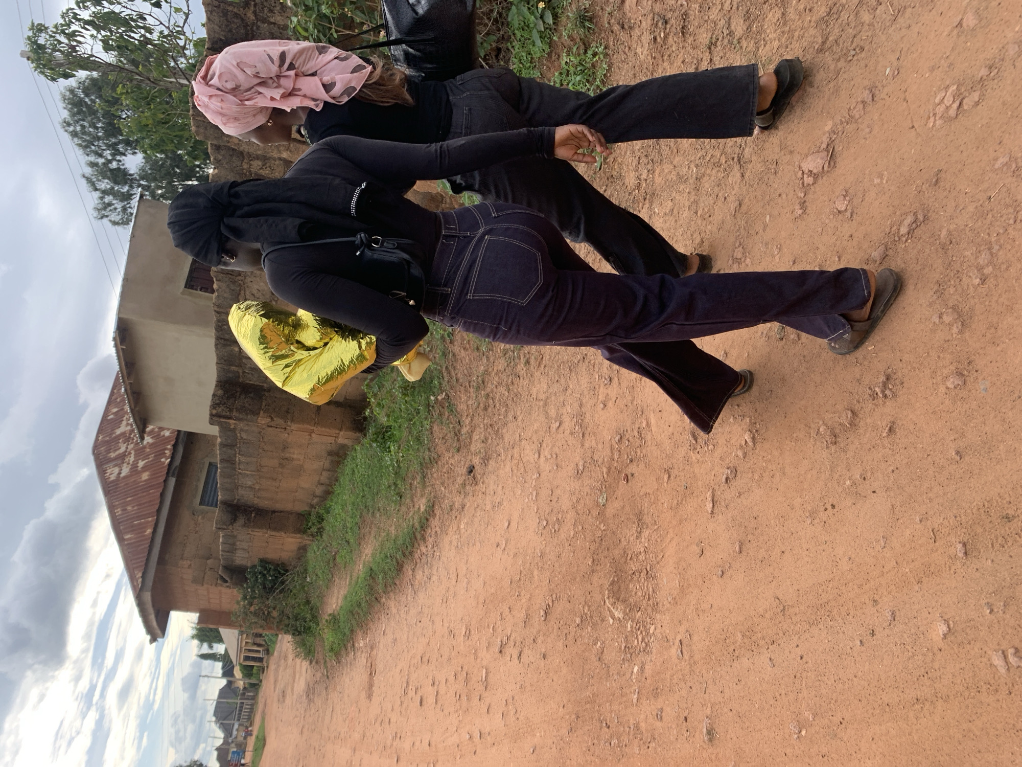 The image depicts two individuals, seen from behind, walking away from the camera on a reddish-brown dirt road in Jos, Nigeria, under an overcast sky.
The person on the left, closer to the camera, wears a black long-sleeved top, dark blue jeans with prominent light stitching, and dark flat sandals. Her hair is covered by a black headwrap, tied at the nape of her neck. She carries a small black shoulder bag on her left side and holds a large, reflective gold-colored Mylar balloon in her left arm.
The person on the right also wears a black long-sleeved top, dark jeans, and dark flat sandals. Her hair is covered by a light pink patterned headwrap. She carries a black shoulder bag on her right side. Both individuals are walking in the same direction along the unpaved road.
The ground is composed of reddish-brown soil, showing unevenness, small stones, and subtle depressions. A narrow strip of green grass and weeds runs along the right edge of the dirt road, bordering a low wall constructed from reddish-brown concrete blocks. Behind this wall, a single-story building with a rusty, corrugated metal roof and reddish-brown walls is visible. In the background, further along the road, more residential structures are partially visible. Taller trees with green foliage rise above the buildings and horizon. The sky is largely covered with grey and white clouds.