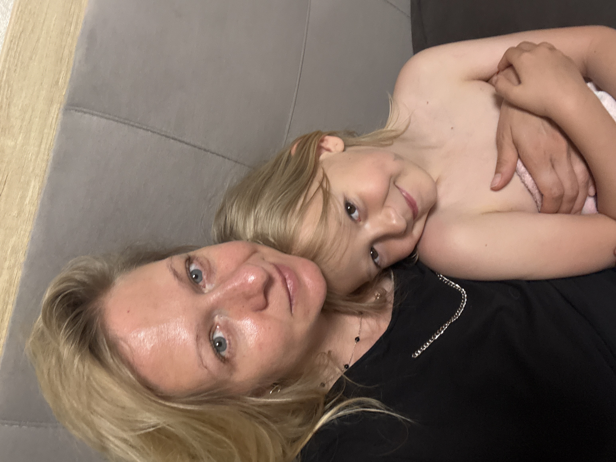A close-up, low-angle shot features an adult and a child lying on a grey sofa. The adult, presumably a woman, has blonde hair, light skin, and blue eyes. She is wearing a black top and a necklace. The child, also fair-skinned with blonde hair, is positioned near the woman's chest. The child is hugging their hands and appears to be wrapped in a pink towel. The background is blurred, indicating a shallow depth of field. The location appears to be an indoor residential space.