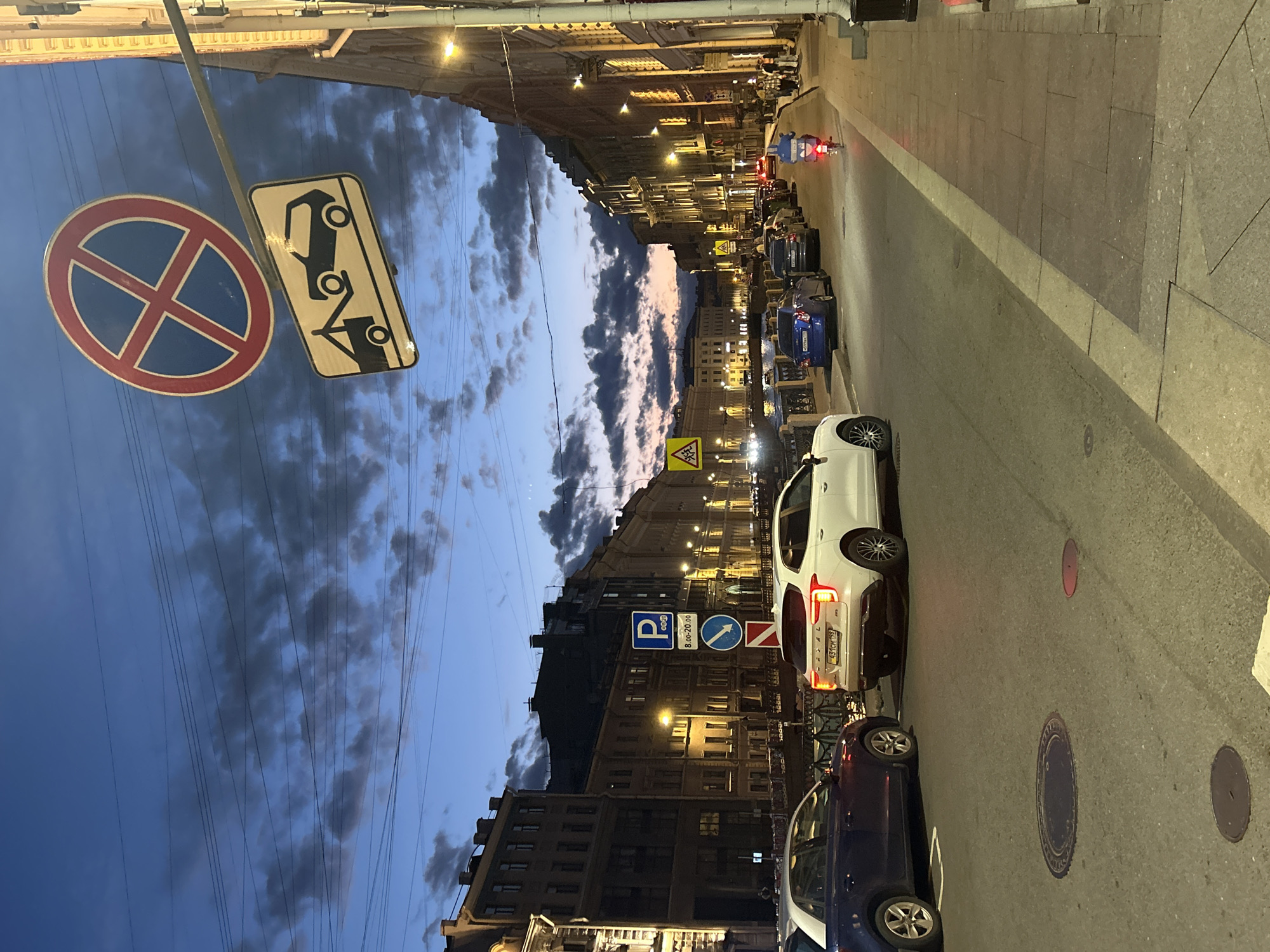 The image depicts a street scene at dusk or twilight, with the sky displaying a mix of blue and dark clouds, hinting at evening or impending weather. The street is lined with multi-story buildings, their windows illuminated, suggesting an urban environment. Several cars are parked along the street, including a white sedan and a dark blue sedan, with their brake lights on. A motorcycle with illuminated headlights is also visible, appearing to be in motion.
Several traffic signs are present. On the left, there is a circular sign with a red border and a blue interior, featuring a red cross, which typically signifies "no parking" or "no stopping." Next to it, a rectangular sign shows a black silhouette of a car being towed. Further down the street, a blue square sign with a white "P" indicates parking, with specific hours of operation (8:00-20:00) noted below. There are also arrow signs indicating direction and a yellow triangular sign with a black symbol, possibly a warning.
The street is paved with cobblestones and asphalt. The overall mood is calm, with the artificial lights of the buildings and vehicles contrasting with the fading natural light. The presence of traffic signs and parked cars suggests this is a public street with regular vehicle traffic. The time of day appears to be late evening, given the dim natural light and the prevalence of artificial illumination. There are no visible people in a prominent way, though there might be figures in the distance or inside buildings. Text on the license plates of the cars is present but illegible.