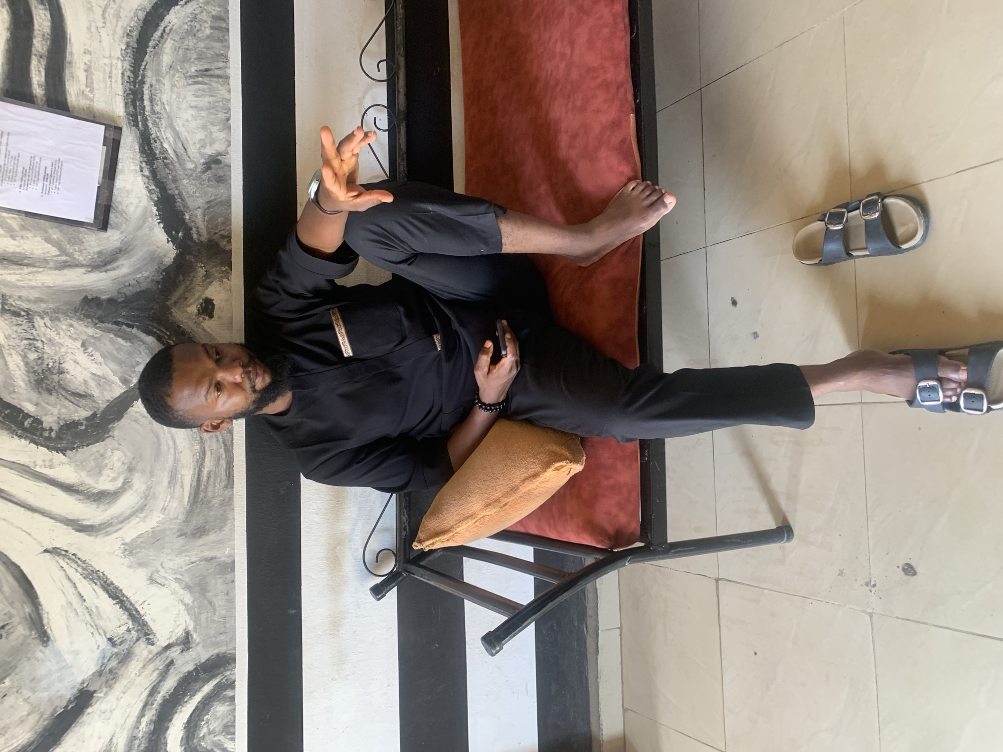 A man reclines on a metal bench with a reddish-brown cushion in what appears to be an indoor setting in Jos, Nigeria. He is dressed in a black long-sleeved tunic with subtle gold-toned detailing on the front placket, and matching black trousers. He wears a watch on his left wrist and a black beaded bracelet on his right. His right arm is bent with his palm open and raised, as if gesturing or speaking. His left hand rests on his abdomen, holding a small, light-brown, textured pillow or cushion. He is barefoot, with his right leg slightly bent and his left leg extended.
The background wall behind him features a large, abstract mural or painting in shades of black, white, and grey, characterized by swirling and broad brushstrokes. To the left, a framed document with illegible text is mounted on this wall. The floor is covered with light-colored, square tiles. A pair of dark grey sandals with light soles and silver buckles is placed on the tiled floor near his feet. The lighting suggests it is daytime. The man has a beard and is looking towards the viewer with a neutral expression.