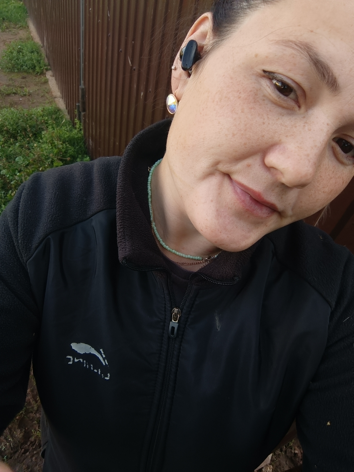A woman with freckles is taking a selfie outdoors. She is wearing a black jacket with a zipper and a logo on the left side of the chest. She has a black wireless earbud in her left ear and a circular, iridescent earring in her left earlobe. She is also wearing a light green beaded necklace. The background consists of a corrugated metal fence and some green grass. The image is taken from a close-up perspective, focusing on the woman's face and upper body. There is no indication of the city of Karsha, Russia, or any other specific location.