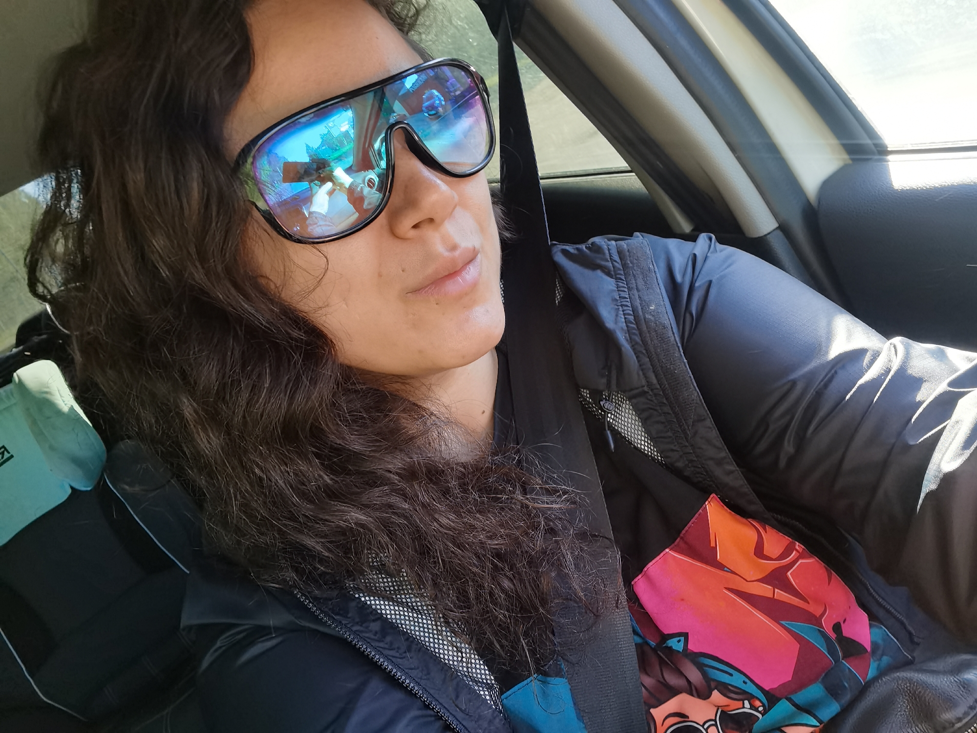 A woman with long, dark, curly hair is taking a selfie from inside a car. She is wearing large, reflective sunglasses that show a distorted reflection of the inside of the car and possibly the person taking the photo. She is wearing a dark jacket over a shirt with a colorful graphic print. A light blue hat is visible on the seat next to her. The background shows the interior of a car, including the window, door panel, and a seatbelt. The lighting suggests it is daytime. This image was taken in Chaikovsky, Russia.