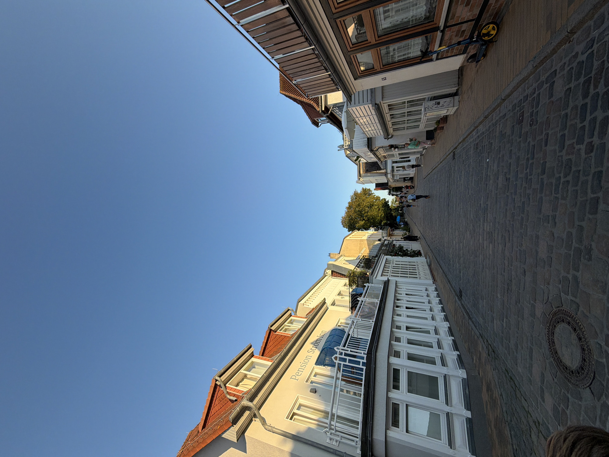 The image depicts a cobblestone street lined with buildings on both sides, under a clear blue sky. The street appears to be in a town or city, with a few people walking along it. On the left side, a building with a sign reading "Pension StromInn" is visible. The architecture suggests a European style, possibly coastal given the presence of beach chairs on a balcony. On the right side, there are more buildings with wooden railings and windows. A scooter is parked near one of the buildings on the right. The overall scene is one of a pleasant, sunny day in a well-maintained urban or town environment. The location has been identified as the Mönchstraße in Rostock, Germany.