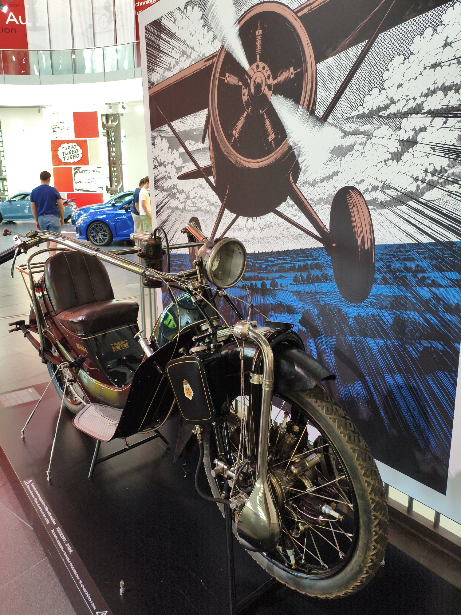 The image is a medium-shot, eye-level photograph taken indoors, likely in a museum or exhibition hall, featuring a vintage motorcycle displayed in the foreground. Behind the motorcycle is a large, stylized illustration of an early airplane in flight, with a blue, abstract landscape below it.
The motorcycle is black and brown, with a prominent leather seat, chrome handlebars, and a large front wheel. It has an old-fashioned headlight and various mechanical components visible. The motorcycle is positioned on a dark, reflective surface.
In the mid-ground, two men and a woman are visible. One man, wearing a blue t-shirt, stands facing away from the camera, looking towards some blue cars parked in the background. Another man, wearing a light-colored shirt, is partially visible next to him. A woman in a light shirt and shorts is also present, standing near the blue cars. The cars appear to be modern sports cars.
The background features a wall with several red and white square panels, some with text and graphics, including one that says "TURBO TURBO TURBO". The overall environment suggests a display related to automotive and aviation history or technology. The context of Ingolstadt, Germany, specifically points to the Audi Museum Mobile, as suggested by the architecture and the presence of Audi cars. The flooring appears to be a dark, polished material.