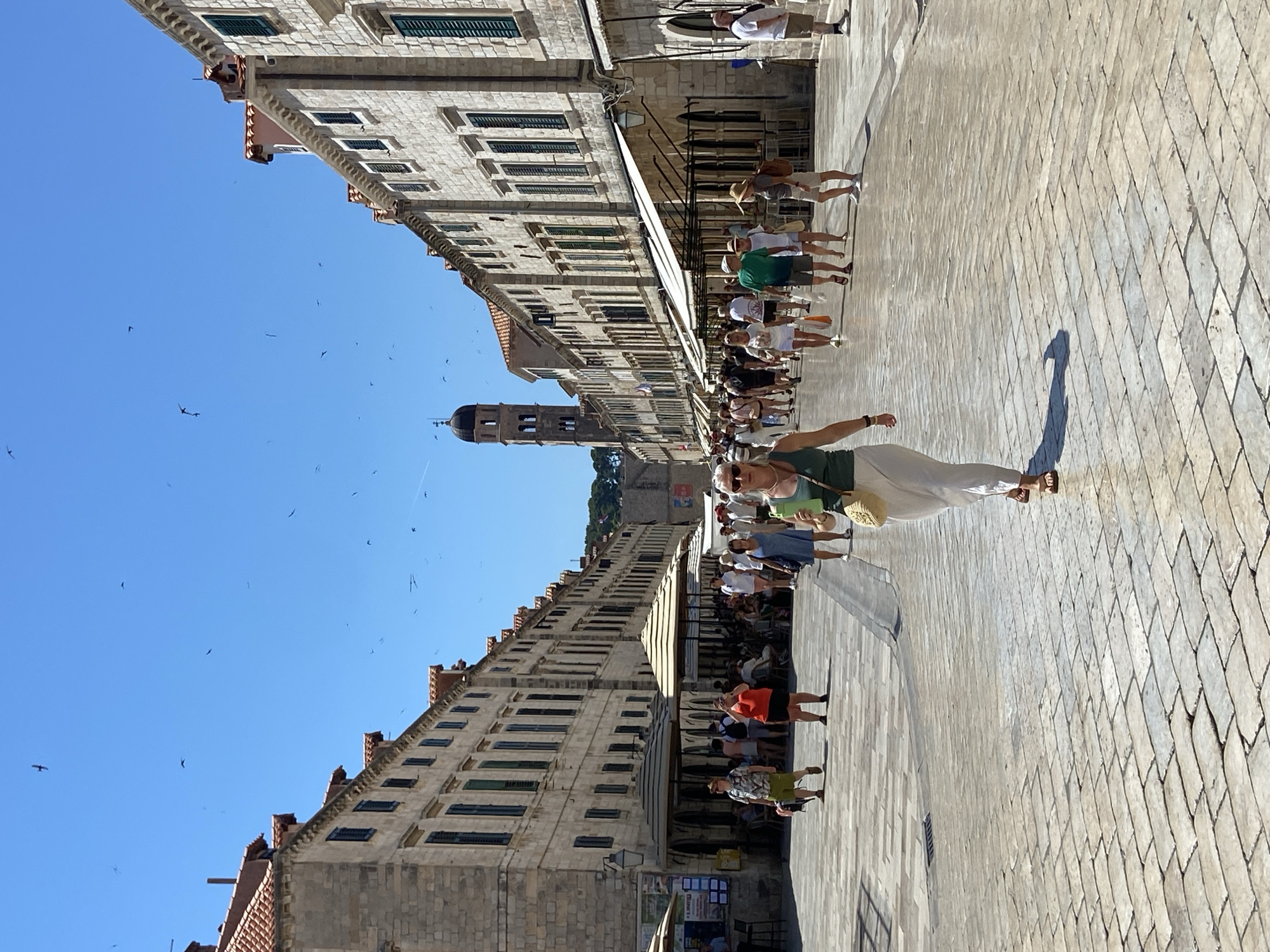 The image captures a sun-drenched, pedestrianized stone street in Dubrovnik, Croatia, likely within the historic Old Town. The street is flanked by tall, stone buildings with characteristic red-tiled roofs and multiple floors of windows, many with green shutters. The architecture suggests a Mediterranean style. Numerous people are walking along the street, some in groups, others individually. Many are dressed in casual summer attire, indicative of tourism. Several outdoor seating areas with umbrellas are visible, suggesting cafes or restaurants. In the background, a prominent bell tower rises above the rooftops, a common feature in European cities. The sky is a clear, bright blue, with several birds in flight. The ground is paved with smooth, light-colored stones. The overall scene conveys a lively, sunny day in a historic urban environment.