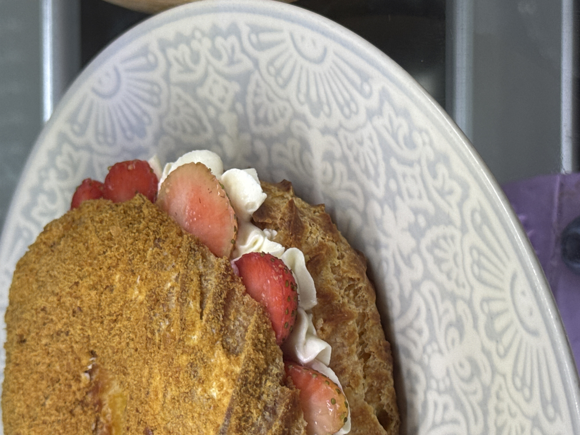 A close-up, eye-level shot showcases a strawberry and cream pastry placed on a patterned plate. The pastry, a circular choux bun, is split horizontally and filled with whipped cream and slices of fresh strawberries. The top bun is coated in a granular, golden-brown topping, possibly crushed biscuits or nuts. The plate features an intricate, light blue floral pattern against a white background. The background is blurred, with hints of gray and darker tones, suggesting an indoor setting, possibly a cafe or bakery counter. There are no people or animals visible in the frame. The overall impression is one of a dessert ready to be served.