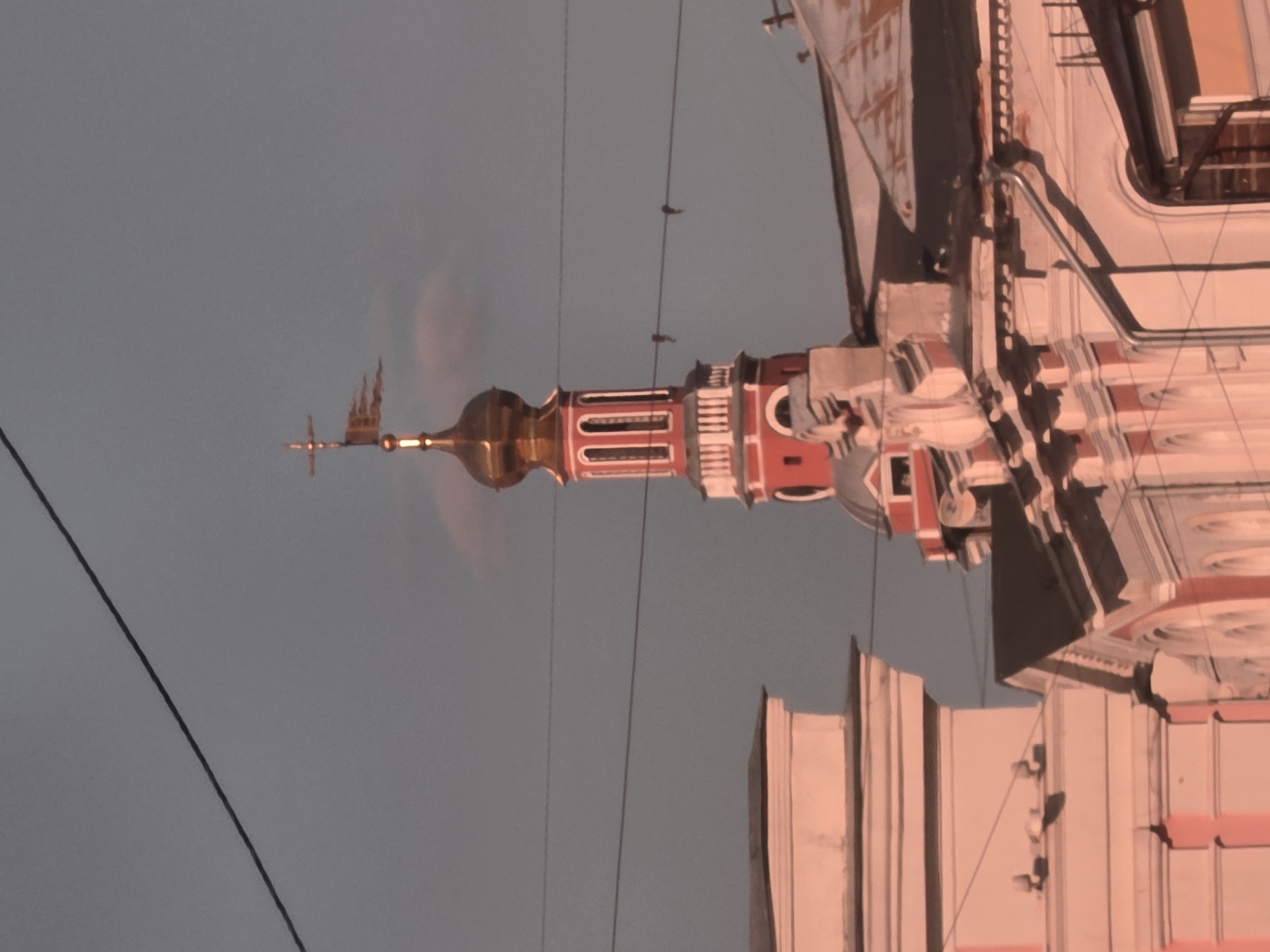 The image shows a close-up, low-angle view of a red and gold onion dome tower with a cross on top, part of a building in Nizhny Novgorod, Russia. The tower is adorned with white trim and arched windows. In the foreground, the upper portions of two adjacent buildings are visible, one with a pink facade and ornate architectural details, the other with a darker roofline. The sky is a hazy gray-blue, with a thin, wispy pink cloud behind the tower. Several dark wires stretch across the frame, obscuring parts of the sky and buildings. The scene appears to be captured during the golden hour, as evidenced by the warm, rosy light bathing the structures. There are no people visible in the image.