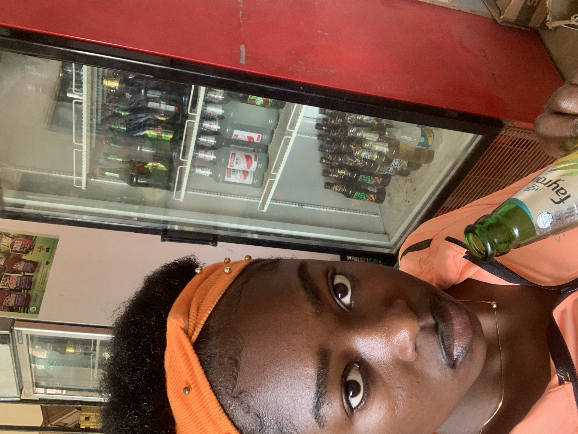 A person is holding a green glass bottle labeled "Fayrouz" in front of a refrigerated display case stocked with various beverages, including bottled beer and soda. The location is plausibly a shop or store in Jos, Nigeria, based on the types of drinks and the general setting. The person is wearing an orange headband with embellishments and a matching orange top. The refrigerated case is red with a black frame.
