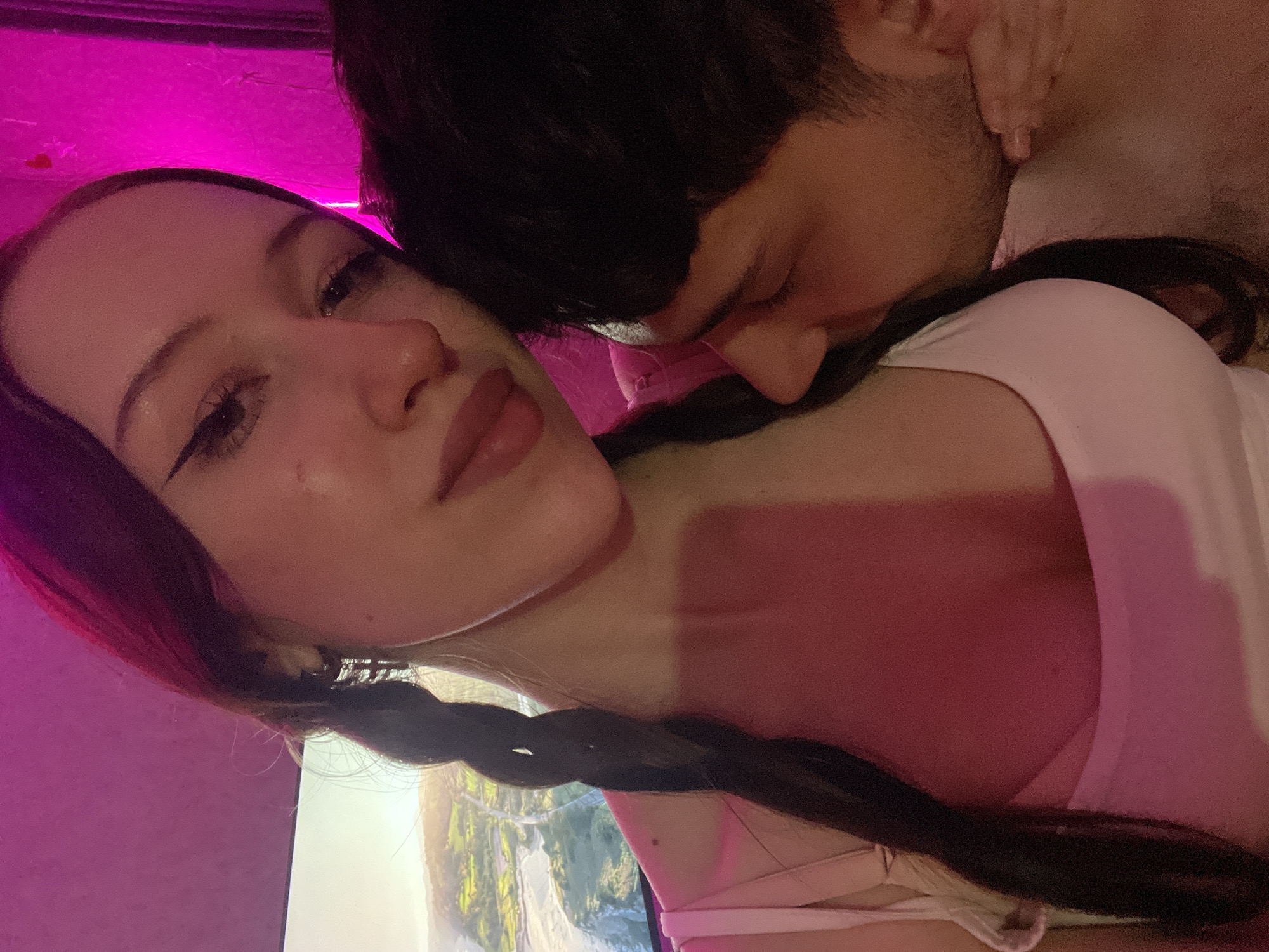 The image depicts an intimate indoor scene featuring a woman and a man, bathed in prominent pink and magenta lighting.
The woman is positioned in the foreground, facing slightly towards the viewer with her head tilted. She has long, dark hair styled in two braids, one of which is clearly visible over her left shoulder. She wears a white tank top with spaghetti straps. Her face is visible, showing dark eyeliner and a soft, direct gaze towards the camera.
The man is situated on the right side of the frame, with his head bent downwards and pressed against the woman's upper chest or neck in an apparent nuzzling or kissing gesture. His face is largely obscured, showing only the top of his head with short dark hair, his nose, and a visible jawline with stubble. A hand, likely his, is partially visible wrapped around the woman's side.
In the background, on the left, a large screen (television or monitor) displays a bright, daytime landscape scene featuring green hills, possibly a body of water, and distant natural formations. The wall on the right side of the frame is intensely illuminated by the magenta light, with faint details like small red heart decorations discernible. The overall environment is dimly lit by artificial colored light, creating an atmospheric setting.