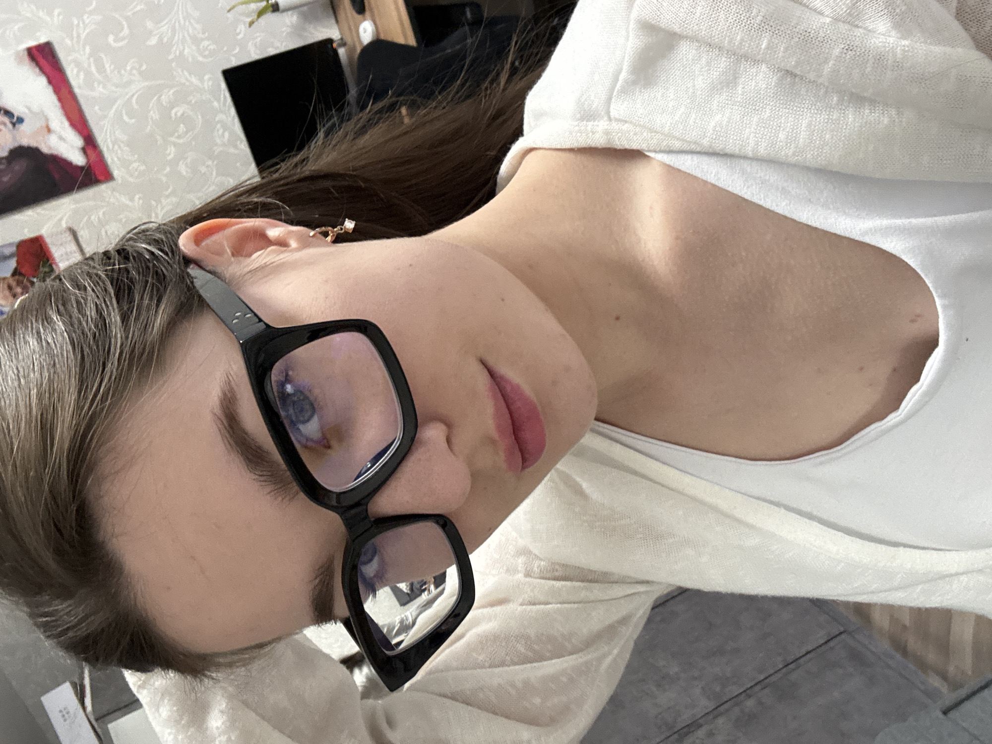 This image is a close-up, front-facing portrait of a female individual, located indoors within the city of Chaikovsky, Russia. The subject has fair skin and dark brown hair, which appears pulled back. She is wearing large, black-framed rectangular eyeglasses, behind which her light-colored eyes are visible. Her lips are naturally colored and slightly parted. She wears a white tank top layered under a loose-knit, off-white or cream-colored open cardigan. A small, gold-toned hoop earring with a clear stone hangs from her right earlobe. Her right hand is partially visible at the upper left of the frame, positioned near her head. The background consists of a wall with light-colored, patterned wallpaper. Two framed pictures are mounted on the wall: one depicting a man against a red background, and another showing a white animal against a red background. To the right, a dark monitor, a dark office chair, a wooden desk, and a small potted plant in a white container are partially visible. The scene is illuminated by ambient indoor lighting.