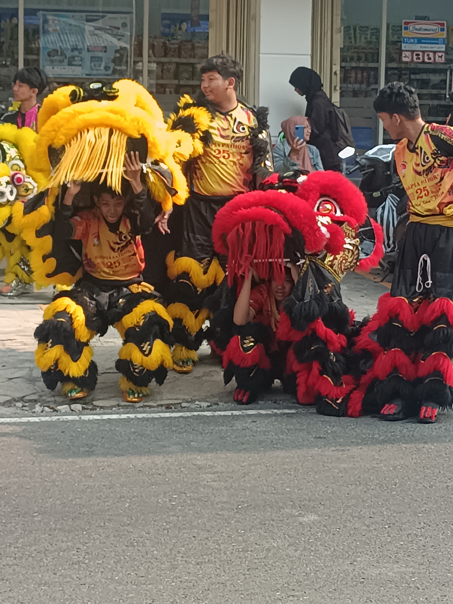 This is a medium shot, taken outdoors on a street in Sleman, Indonesia. In the foreground, two lion dancers are visible, each manipulating the head of a lion costume. The dancer on the left is wearing a yellow and black lion costume with yellow fringes, while the dancer on the right is wearing a red and black lion costume with red fringes. Both dancers are young males and appear to be performing as part of a cultural event. Behind them, other people are gathered, including individuals in matching yellow performance shirts and others observing the event. The background shows a building with a store, indicated by signage such as "Indomaret" and "TUK." The street is paved and has a white line marking.